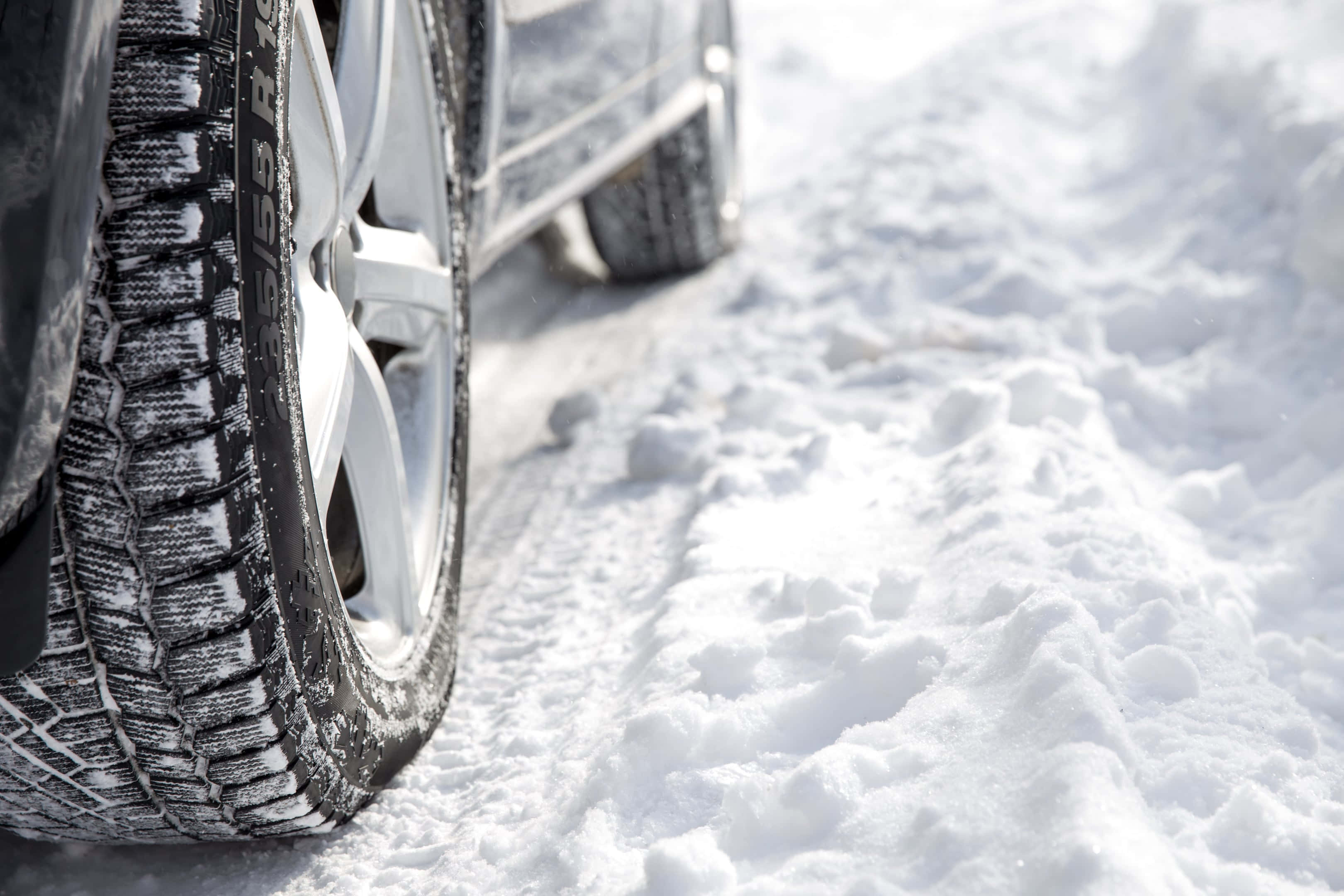 Winter Driving 桌布 壁紙 — 雪地冬季輪胎的特寫，背景有一輛銀色汽車在雪路上。