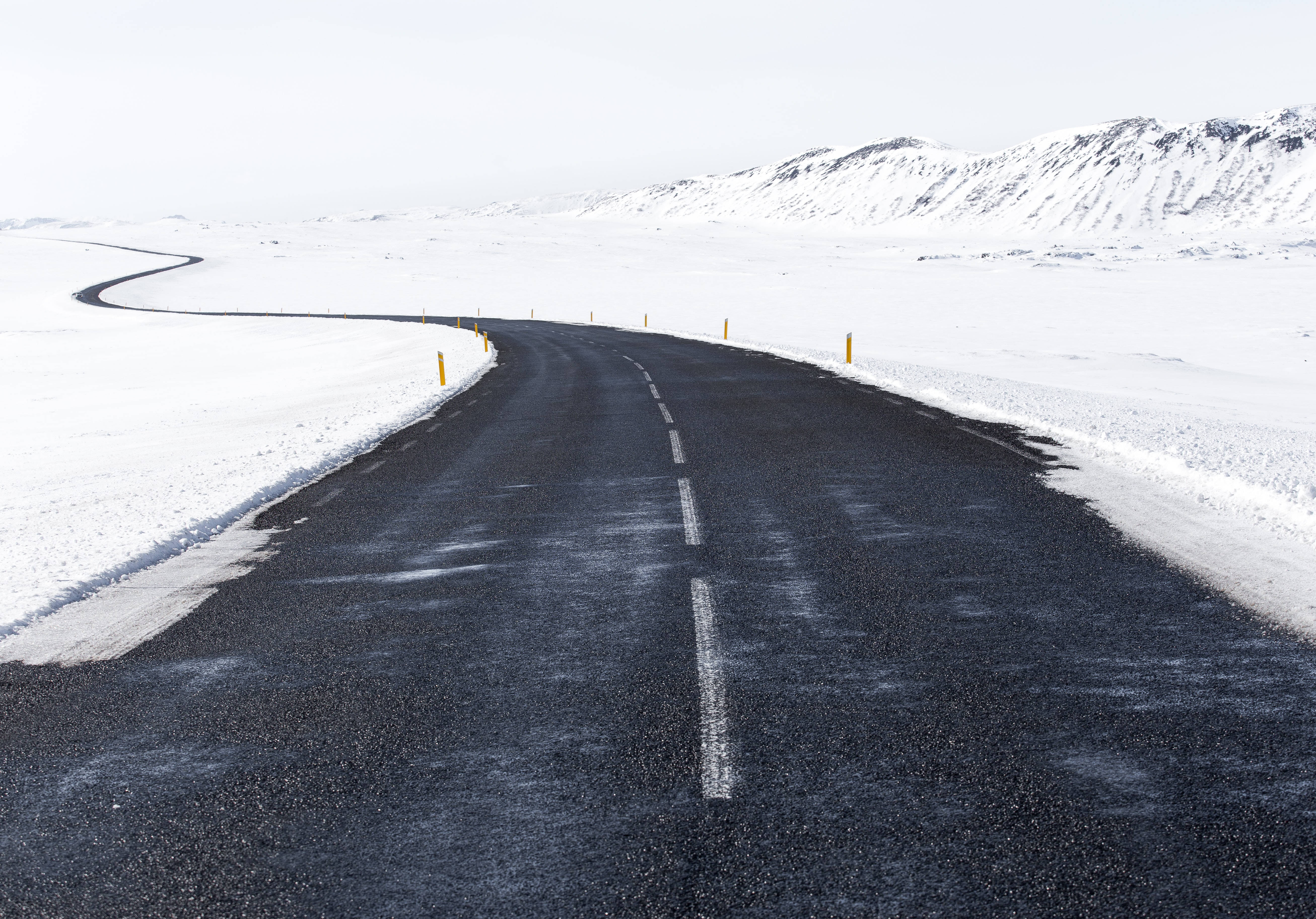 雪地路面Tumblr桌面 壁紙 — 一條蜿蜒的瀝青路穿過廣闊的雪景，遠處有白雪覆蓋的山脈，天空蒼白。