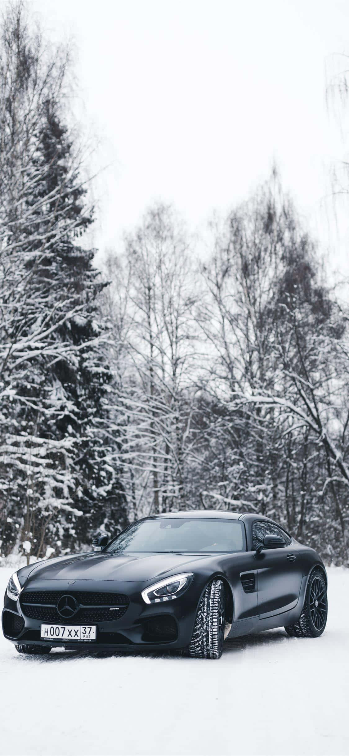 雪地黑色 GT iPhone Xs Mercedes AMG 背景 壁紙 — 一架時尚的黑色Mercedes-AMG GT停泊在白雪覆蓋的森林中，雪覆蓋的樹木在陰天之下。