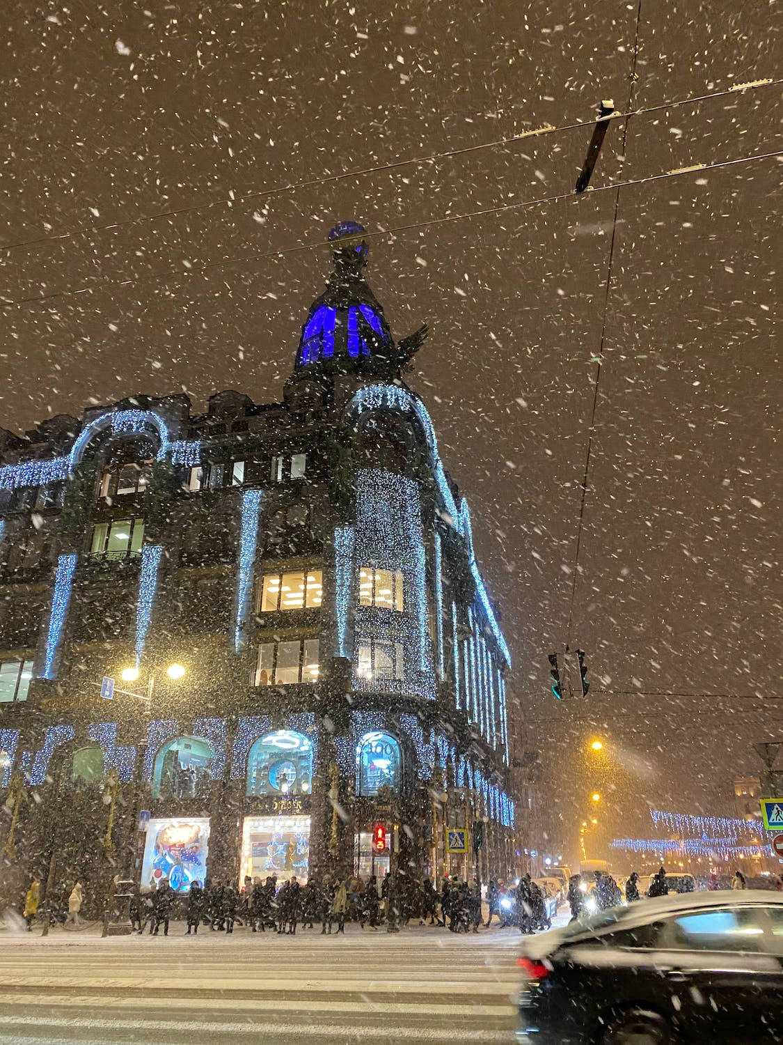 夜間，明亮的城市街道上飄著雪，有行人和汽車。