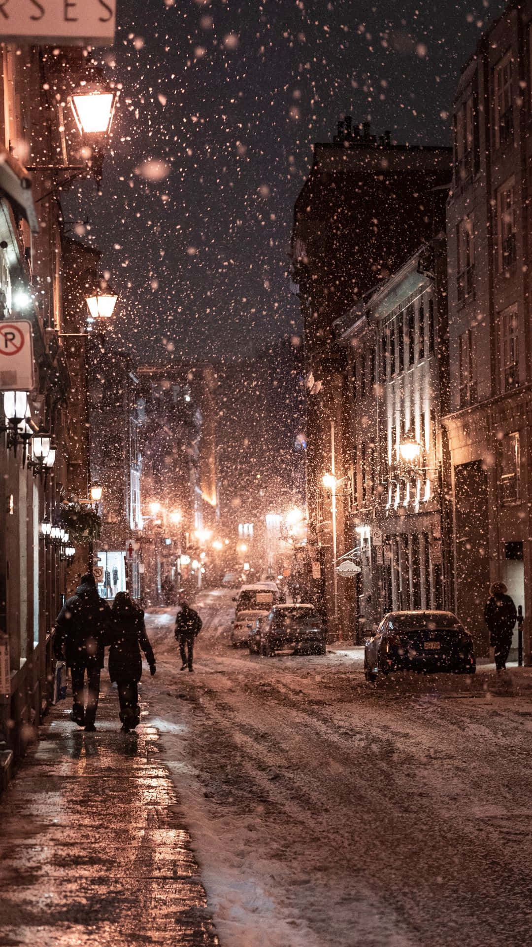 夜間城市街道上飄雪，有發光的街燈和行人。