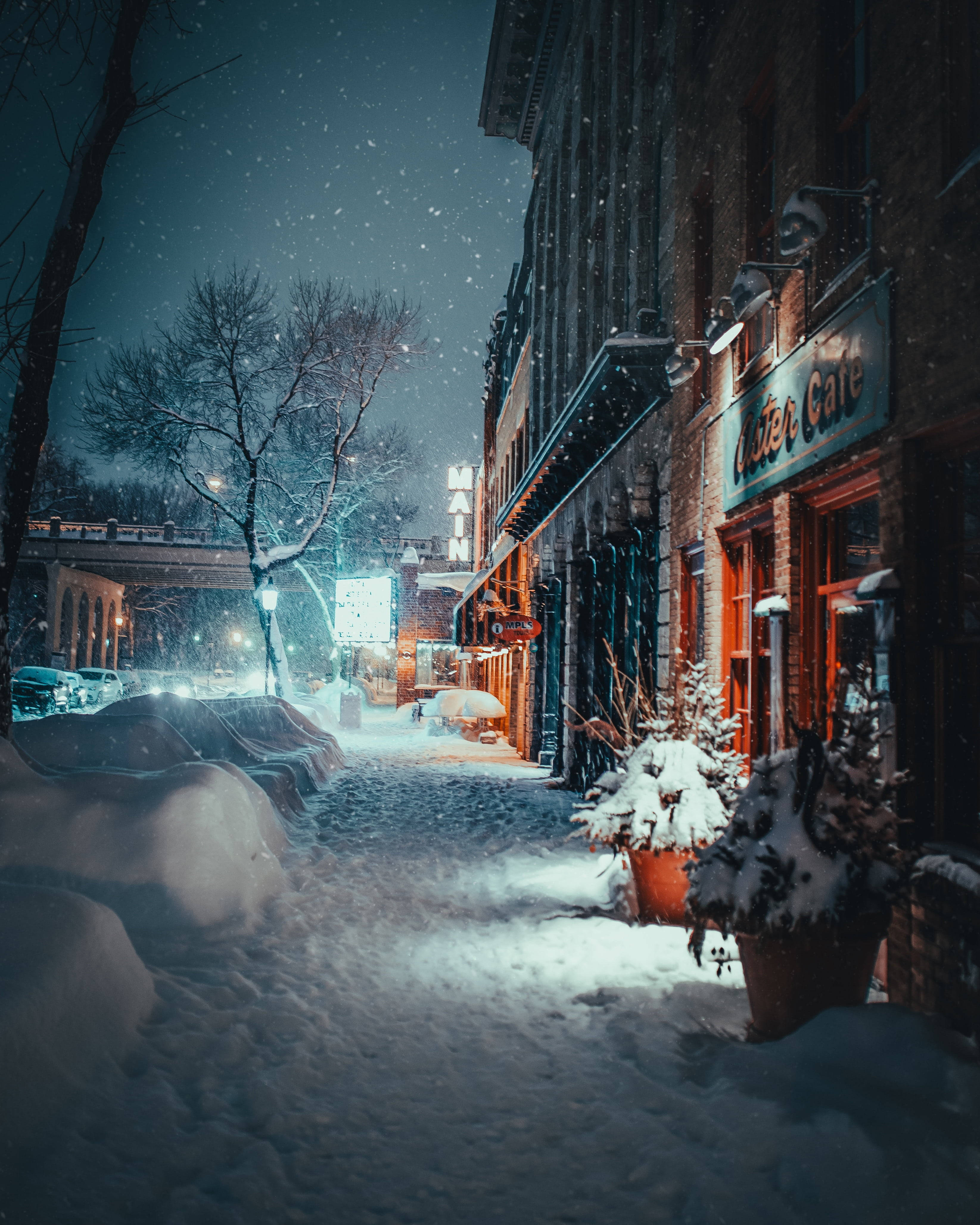 雪夜街角溫馨咖啡店 壁紙 — 白雪覆蓋寧靜的城市街道，夜晚時分，店舖櫥窗的溫暖燈光在飄落的雪花和漆黑的天空中閃耀。