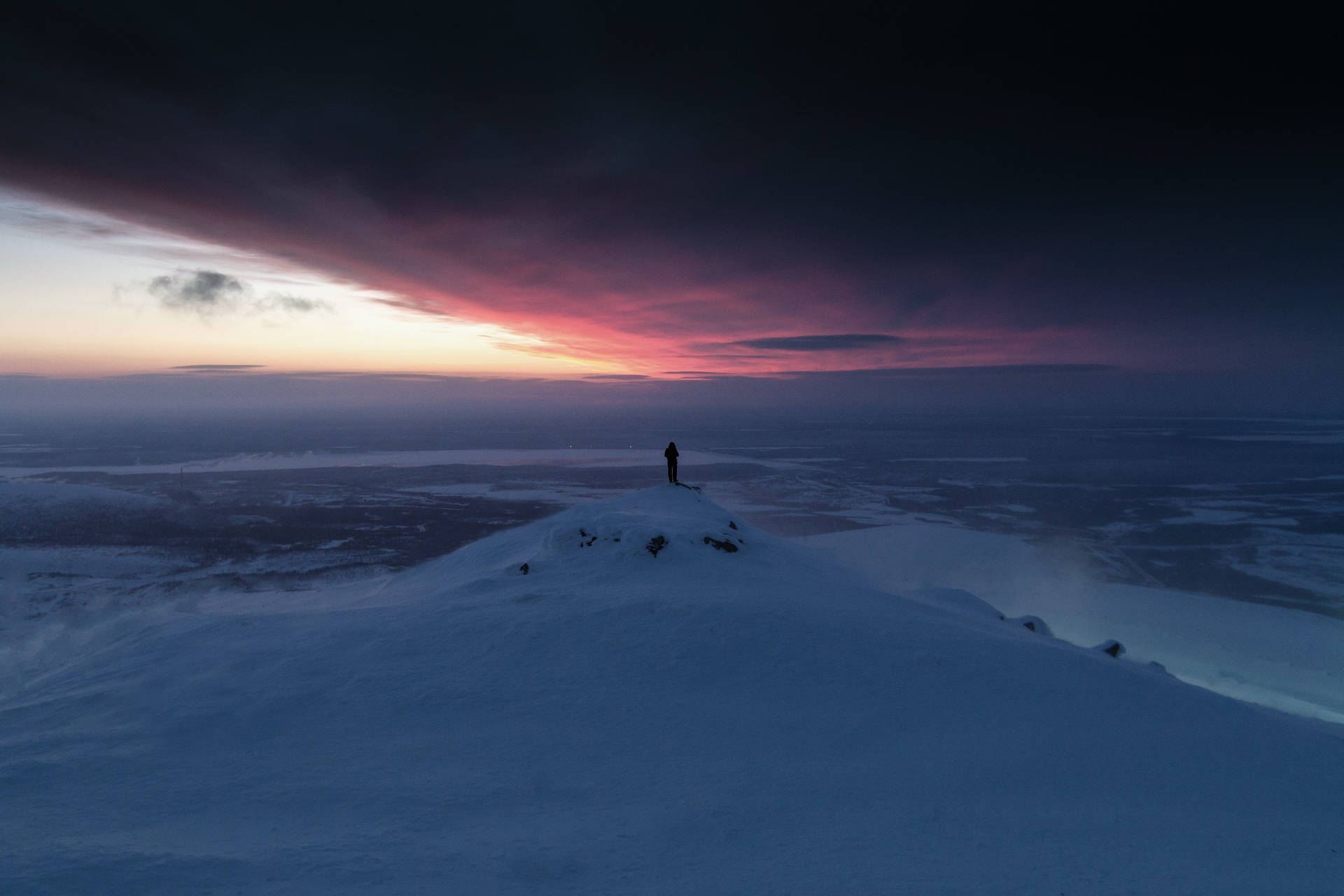 一個人獨自站在白雪覆蓋的山頂，在夕陽下俯瞰冰凍平原。