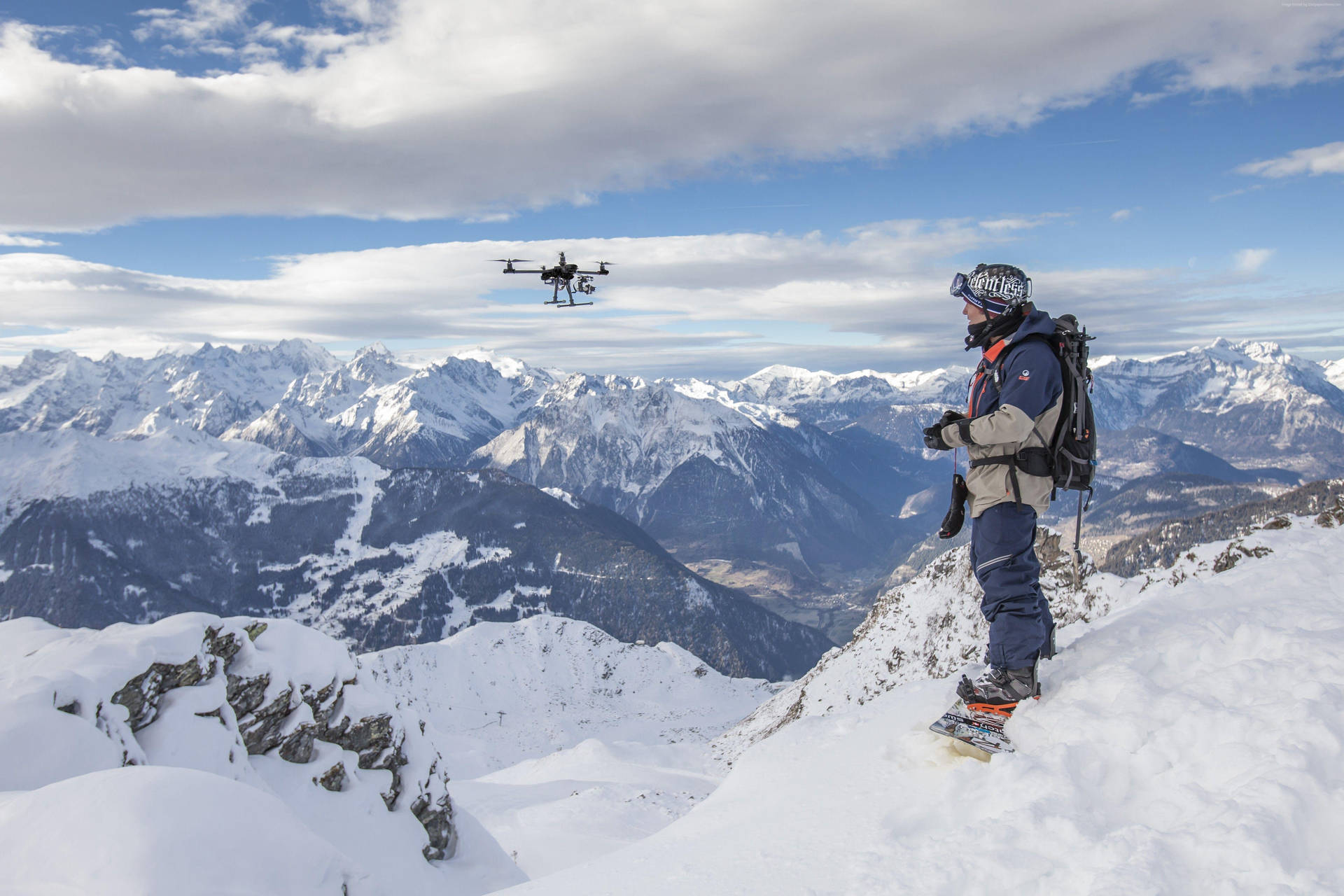 雪山上的 GoPro 攝影登山者 壁紙 — 一名滑雪板手站在白雪覆蓋的山頂上，操控著一架無人機，在部分多雲的天空下盤旋在廣闊的阿爾卑斯山谷之上。