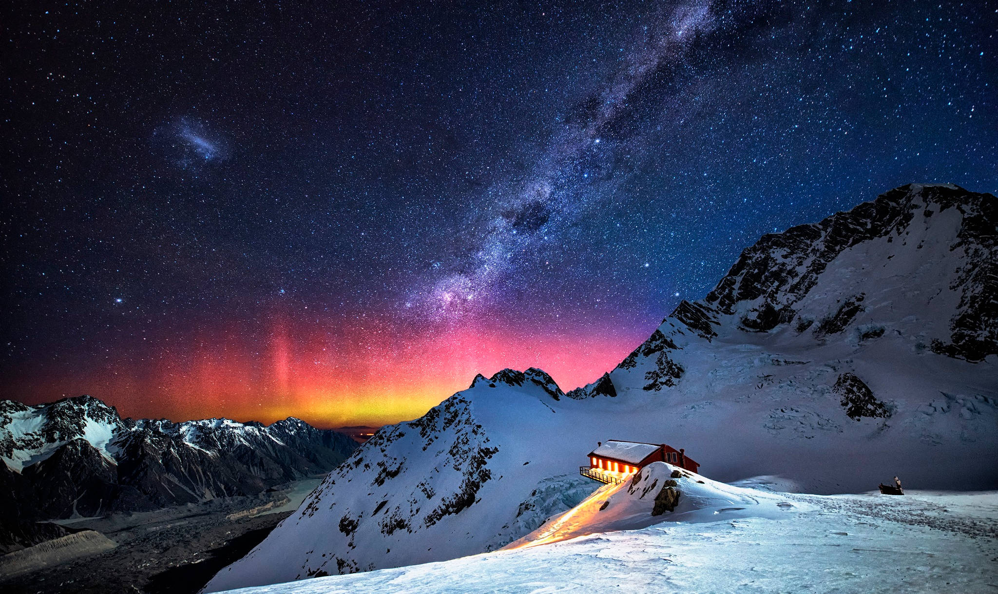 雪山上的星河舞動 壁紙 — 一間舒適的紅色山間小屋在充滿活力的星空下閃耀，北極光拱形橫跨雪覆蓋的山峰。