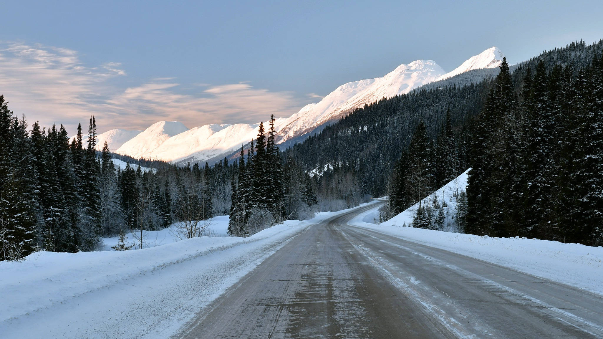 雪山冬季道路 壁紙 — 一條蜿蜒的積雪道路穿過茂密的松樹林，通往日出時雄偉的雪山。