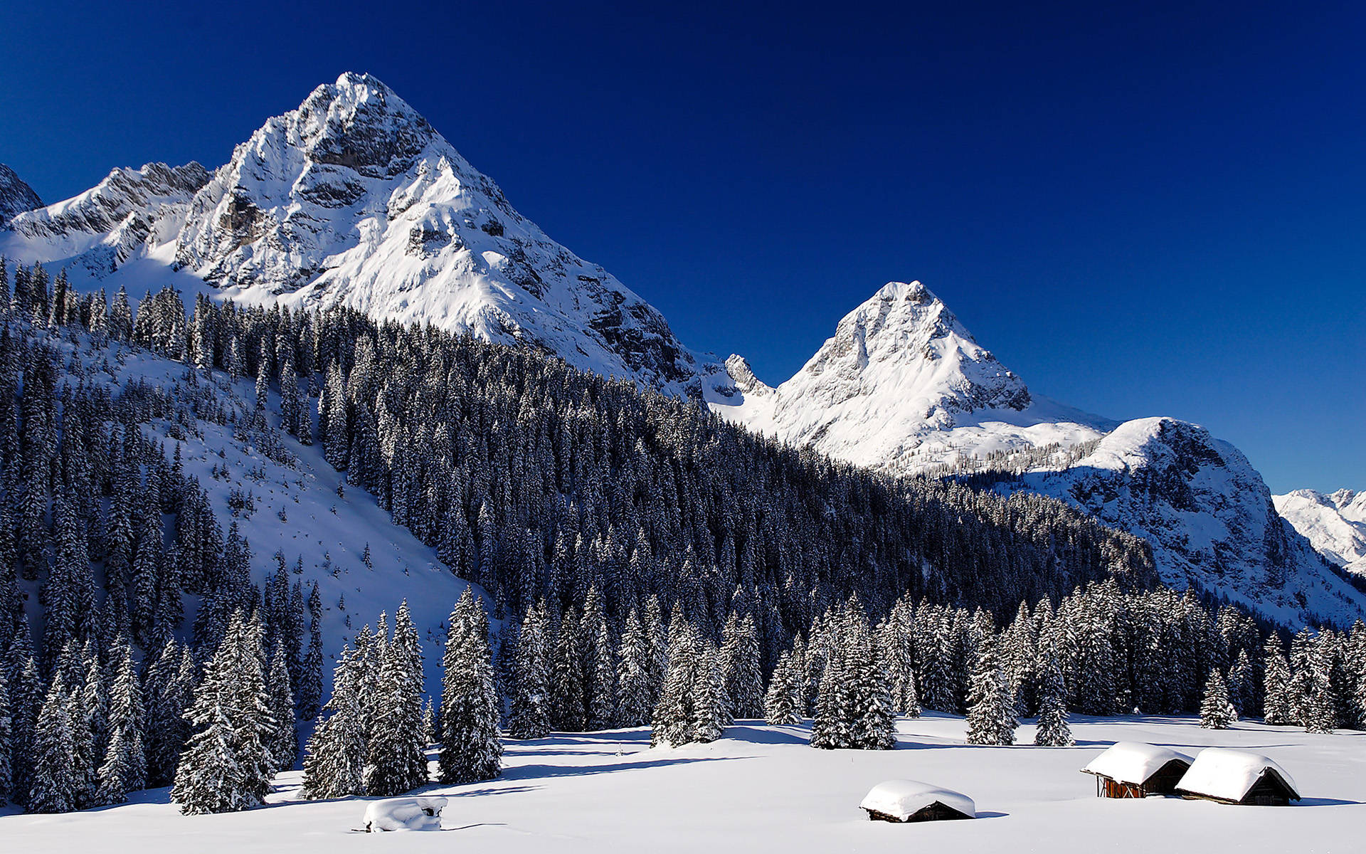 雪山冬季風景 壁紙 — 白雪覆蓋的山峰在深藍色天空下，在一片積雪的松樹和小木屋森林之上，陡峭地聳立。