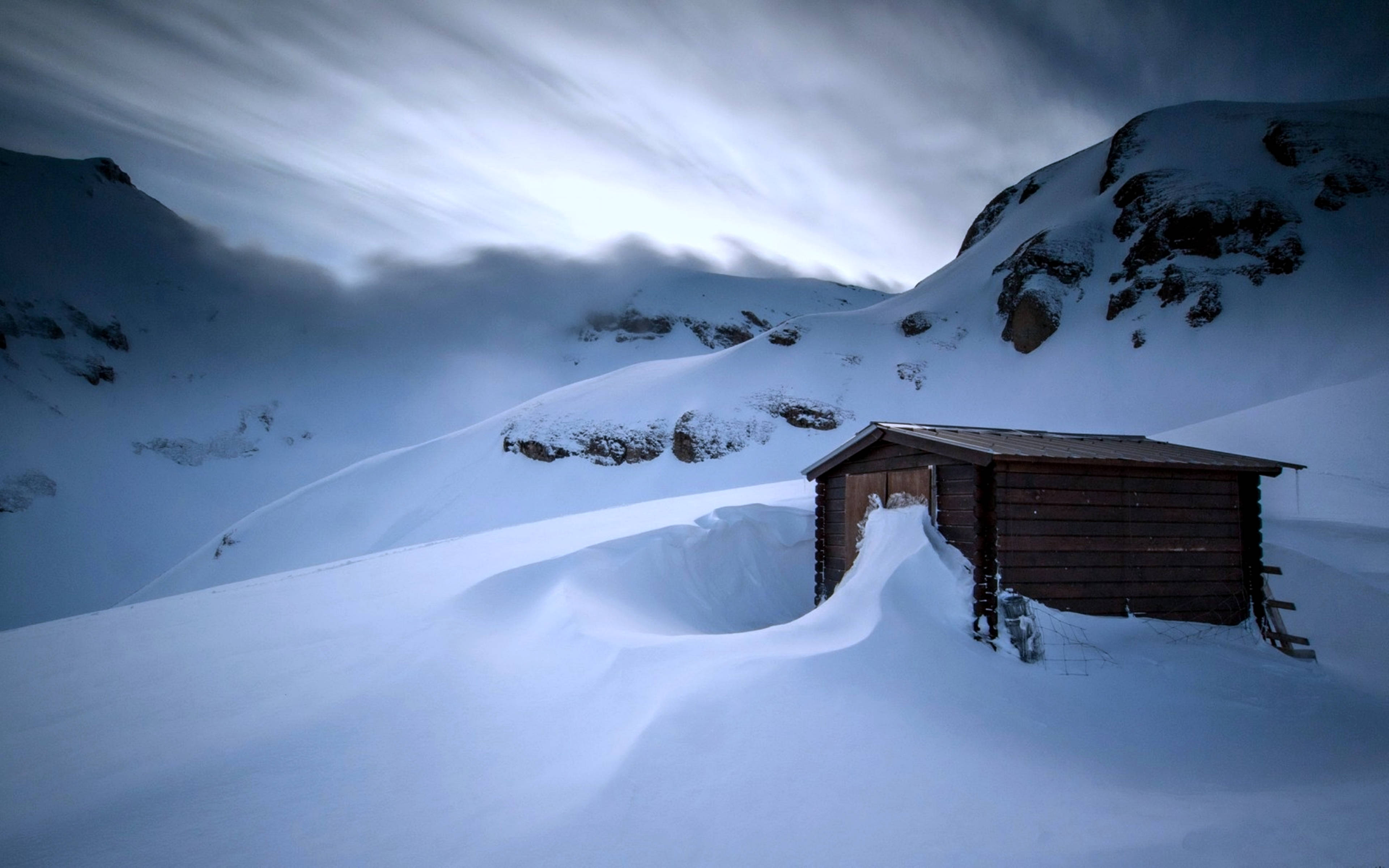 雪山小屋冷景 壁紙 — 在戲劇性的多雲天空下，一座孤獨的木屋埋在深雪中，位於山區冬季景觀中。