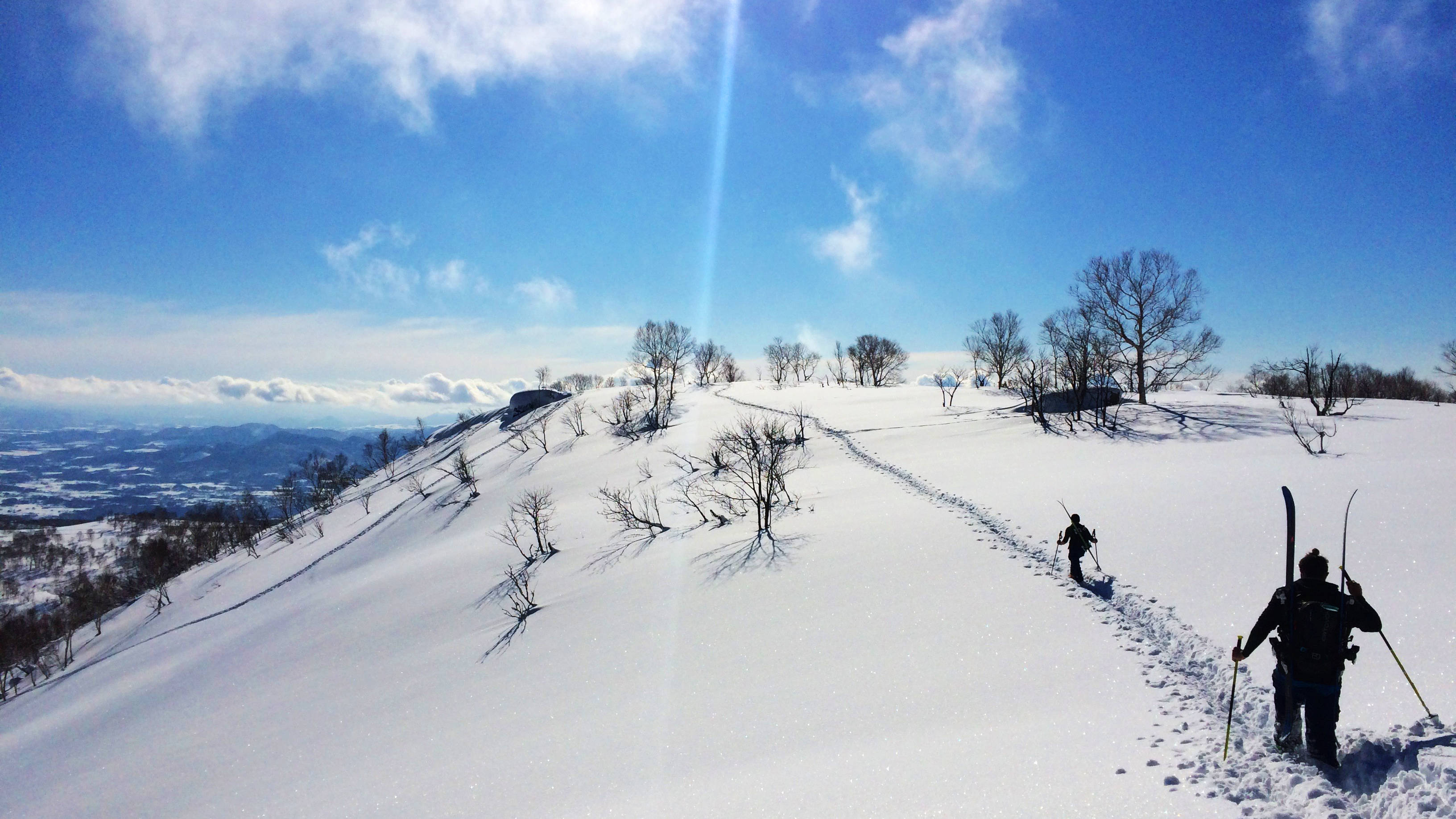 雪山小徑 壁紙 — 兩名滑雪者在明亮的藍天白雲下，沿著積雪的山脊向上跋涉。