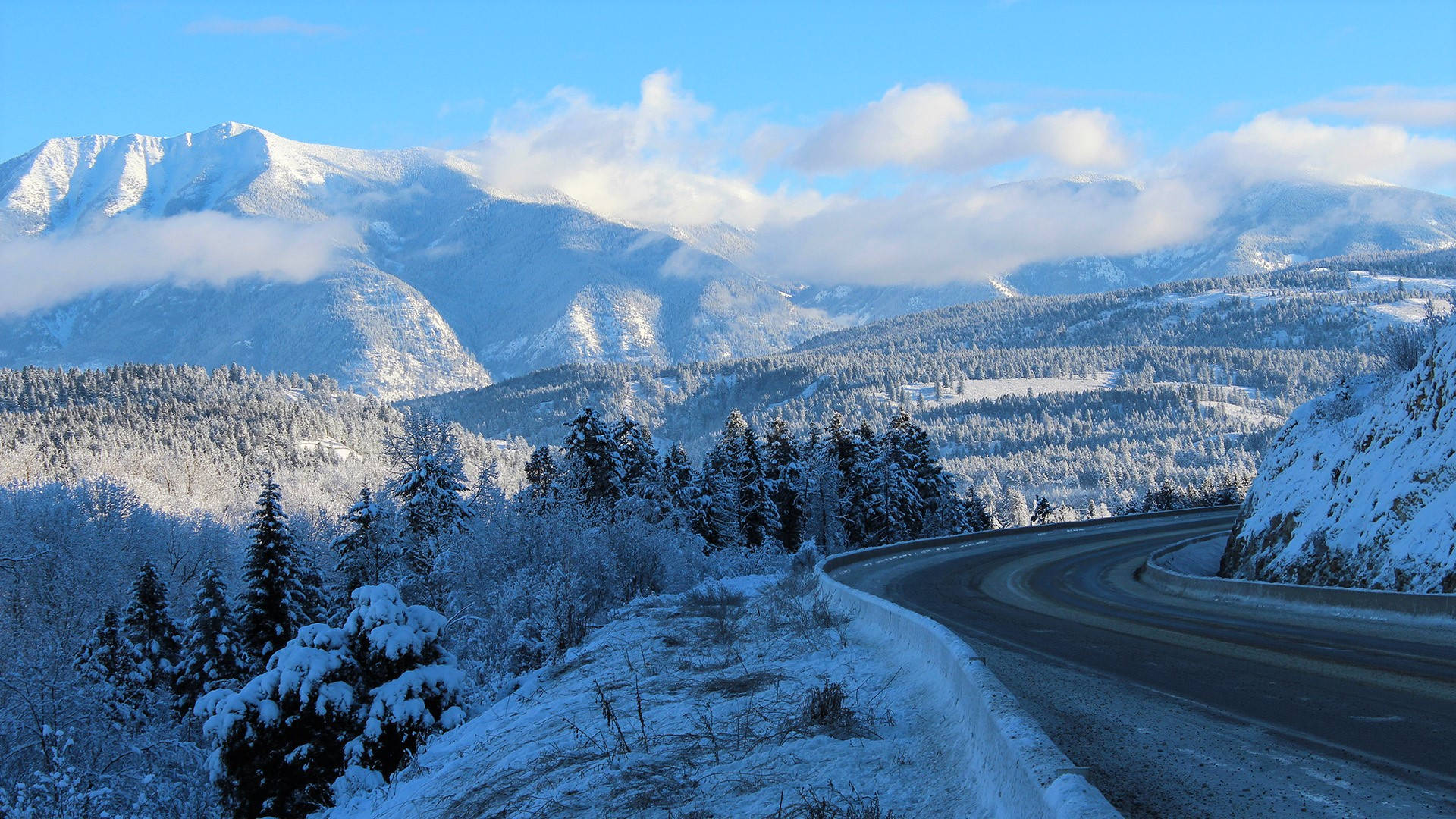 雪山峽谷通道 壁紙 — 一條蜿蜒的道路穿過白雪覆蓋的山脈，在明亮的藍天下有蓬鬆的雲朵。