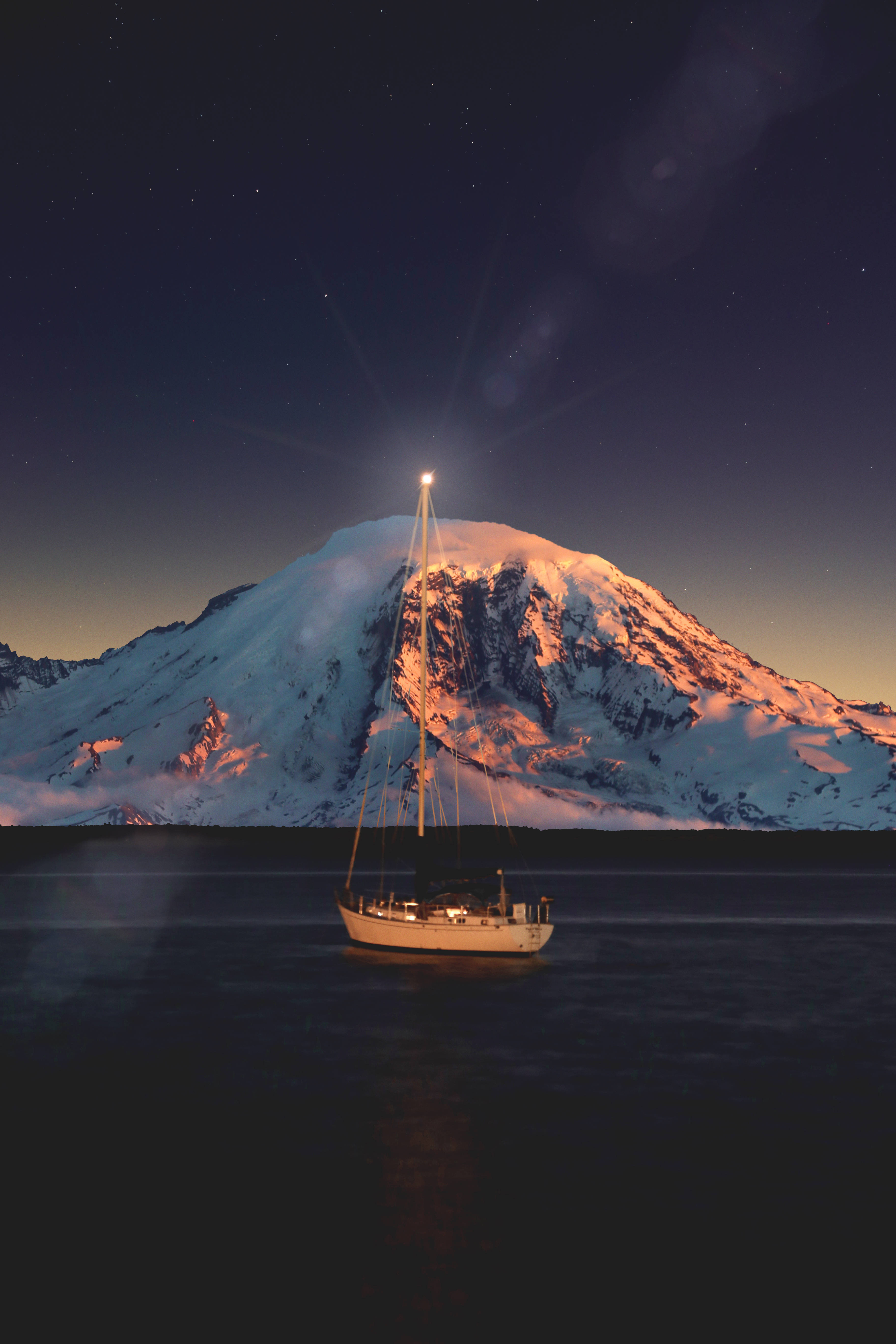 雪山近海航行 壁紙 — 一艘帆船在平靜的水面上溫暖地發光，星空下，一座積雪的山峰在金色暮色中聳立。