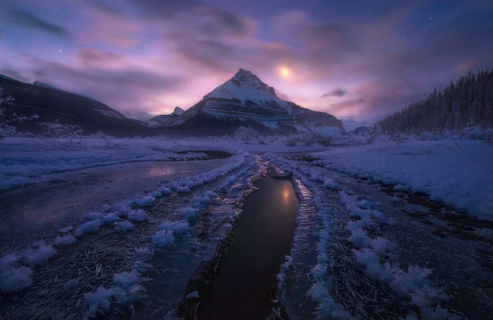 雪山長鏡頭遠景 壁紙 — 一條冰封的河流蜿蜒穿過雪谷，在紫色暮色天空下，一輪發光的月亮懸在積雪的山峰之上。