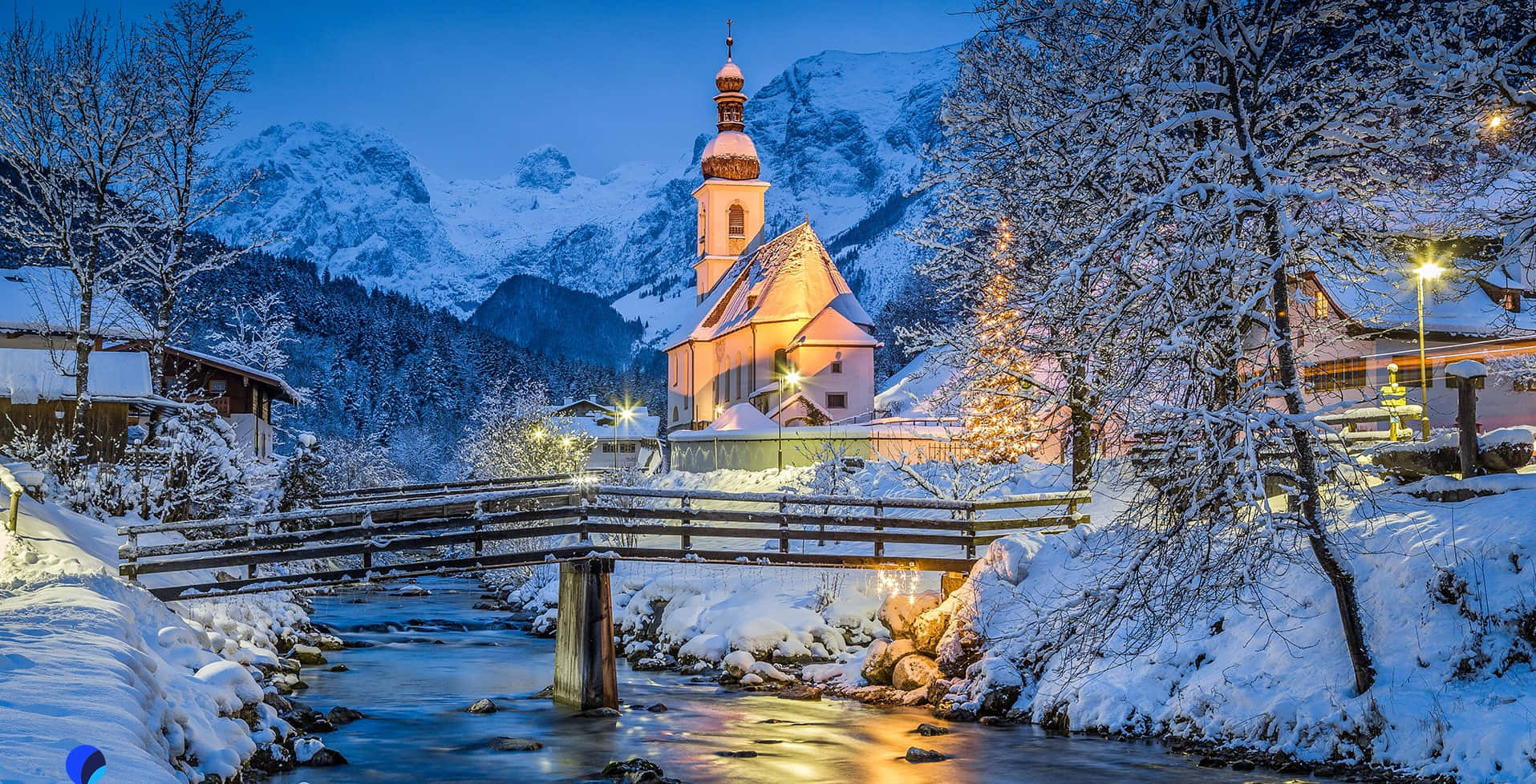 雪山風景，遠眺壯麗山脈 壁紙 — 一座被雪覆蓋的教堂在暮色中溫暖地發光，一座木橋橫跨山村的溪流。