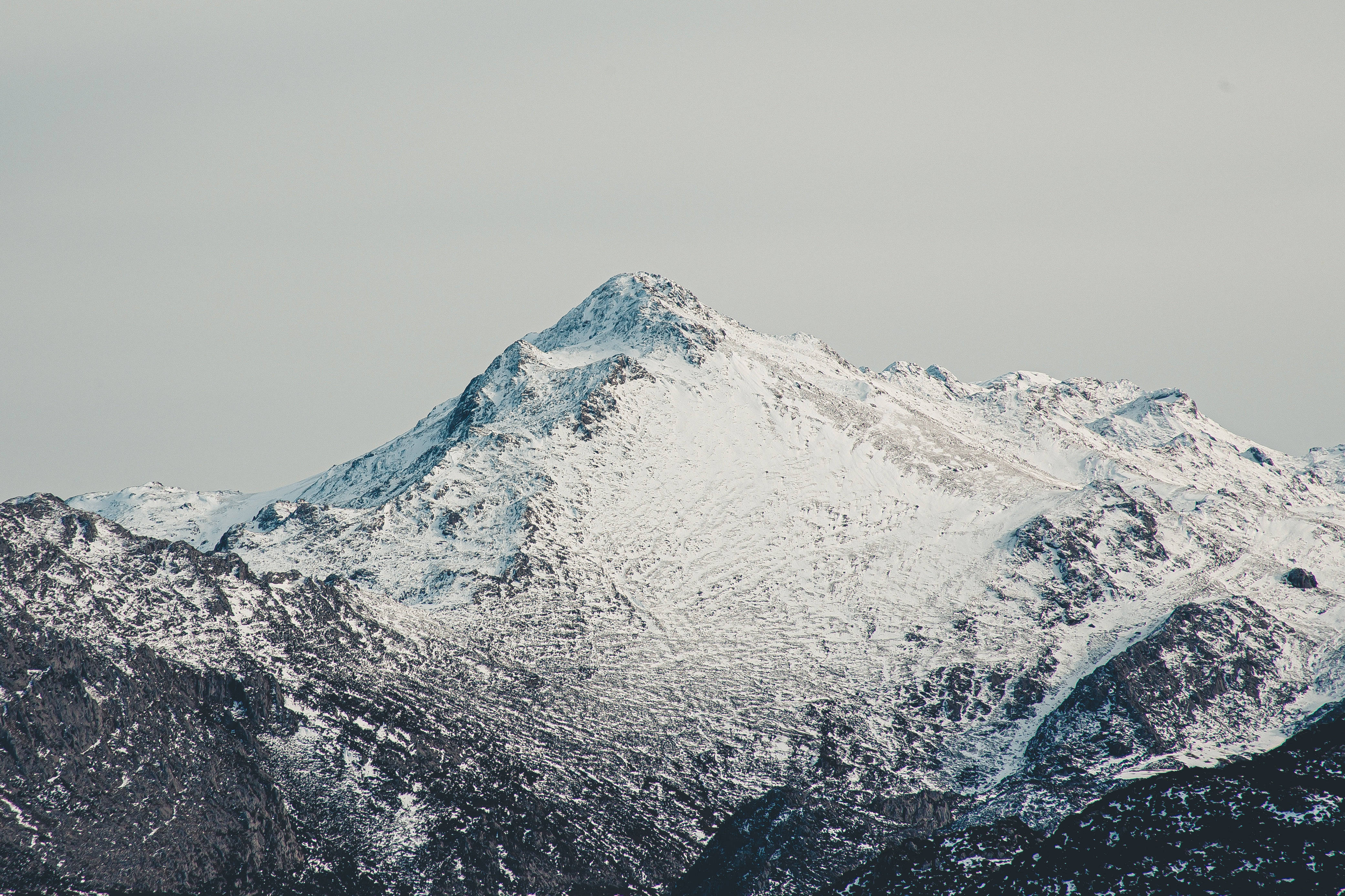 雪峰最美自然風景 壁紙 — 一個積雪的山峰在淡灰色的天空下陡峭地聳立，下方是質感豐富的岩石斜坡。