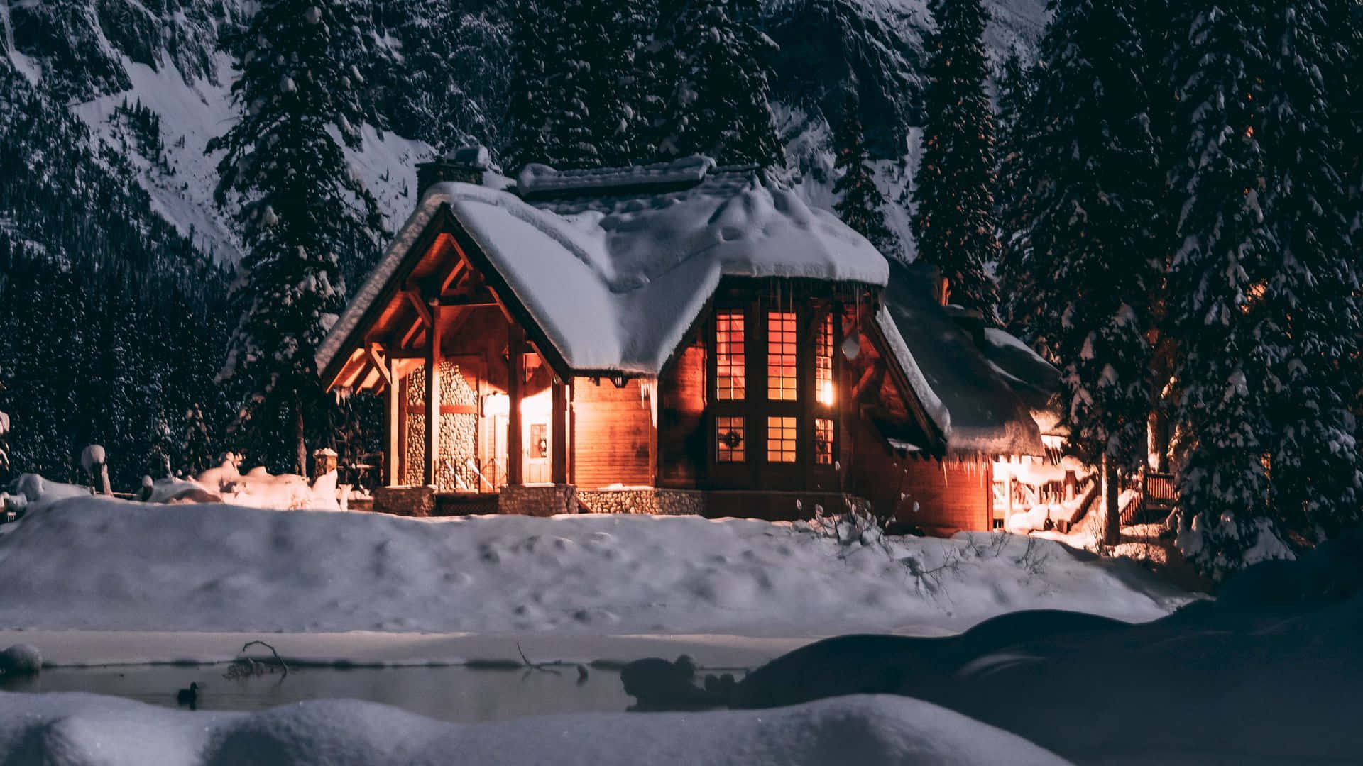 雪景冬季風光 壁紙 — 一間舒適的雪覆蓋木屋在夜晚溫暖地發光，坐落在山區冬季森林中積雪的松樹之間。