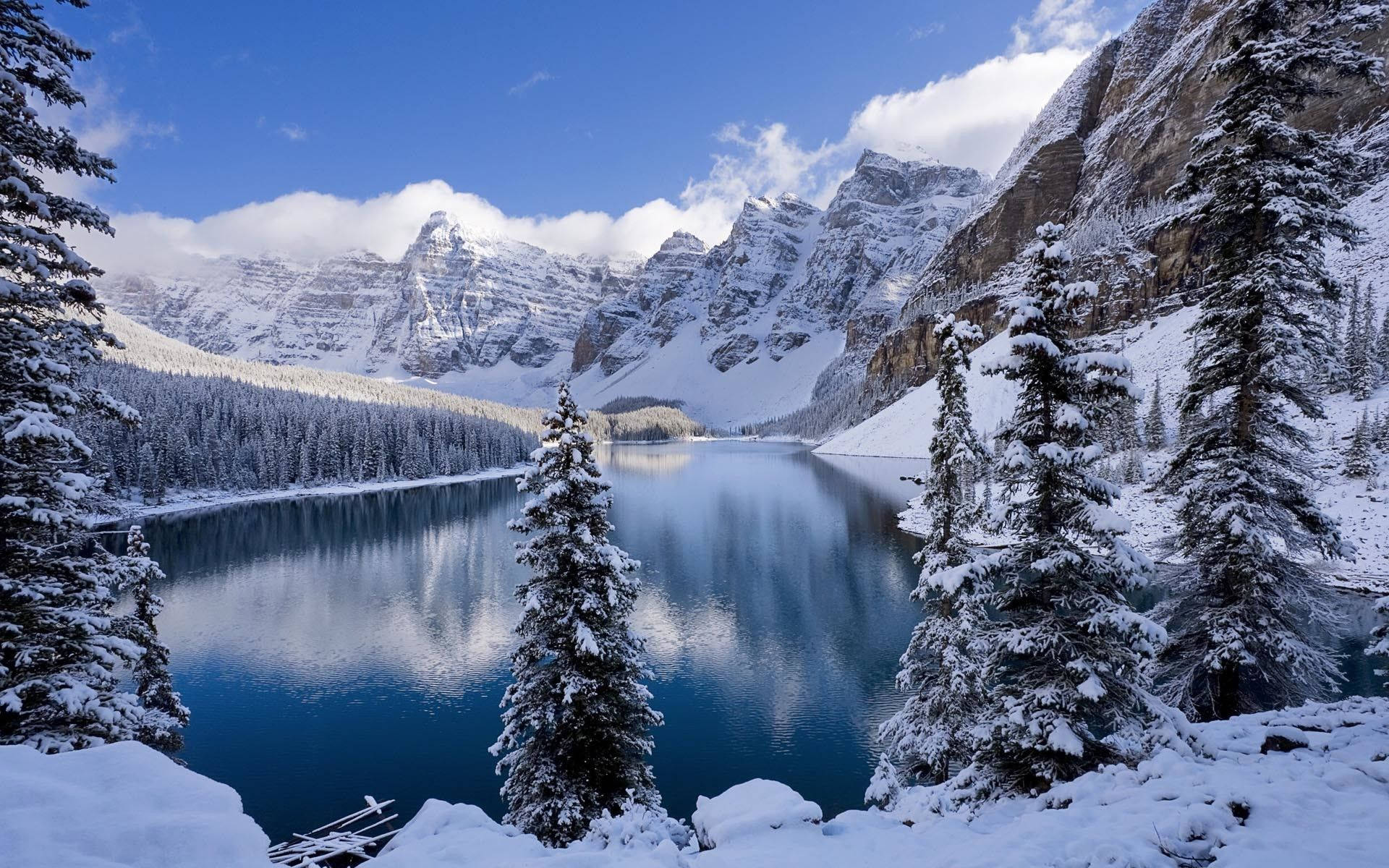 雪景湖畔冬日風光 壁紙 — 寧靜的冬日湖泊映照著白雪覆蓋的山脈和常綠樹木，天空湛藍，雲朵蓬鬆。