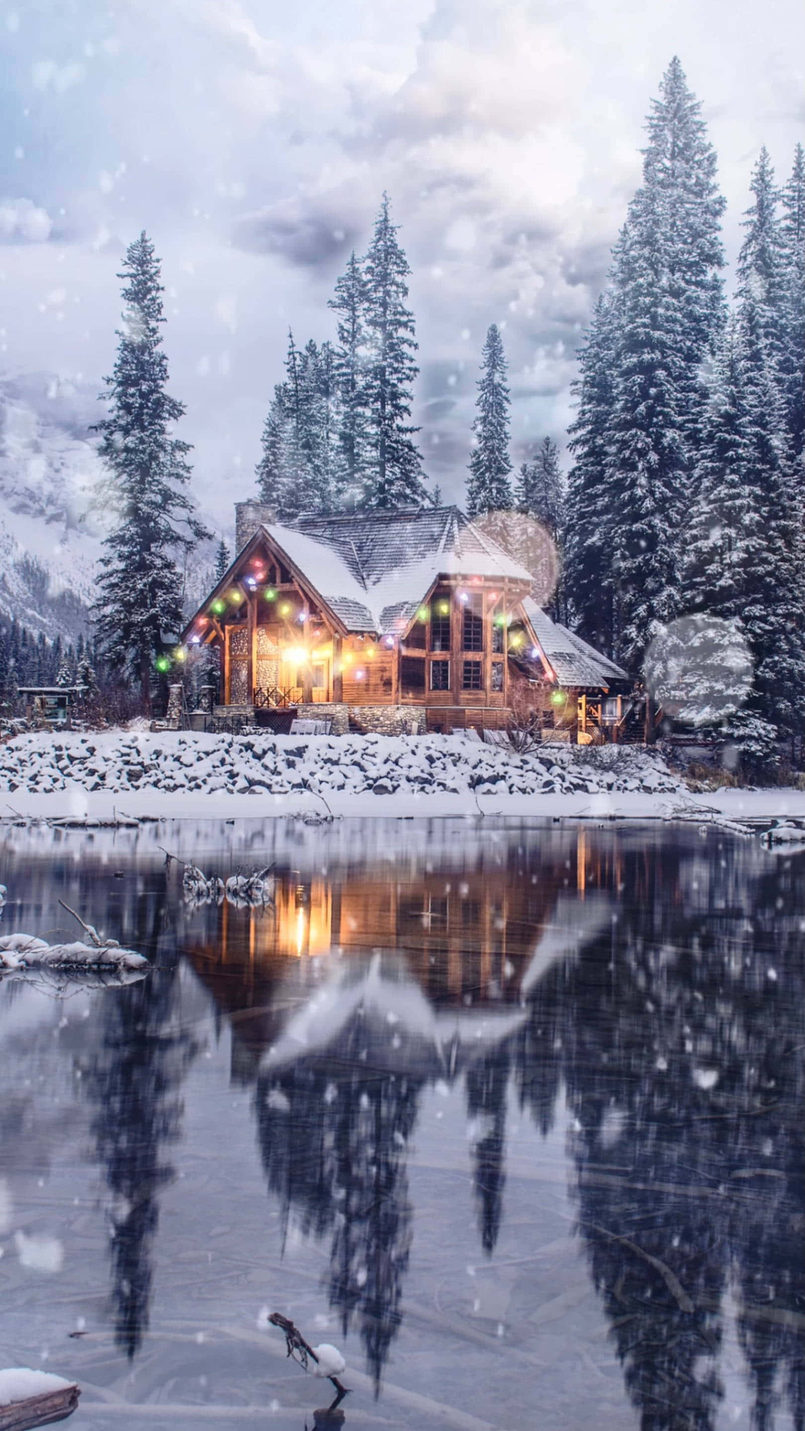 雪林冰湖小屋鏡面攝影 壁紙 — 一間舒適的雪覆蓋小屋，溫暖的燈光在結冰的湖邊閃爍，映照著松樹，背景是雪黃昏的天空。