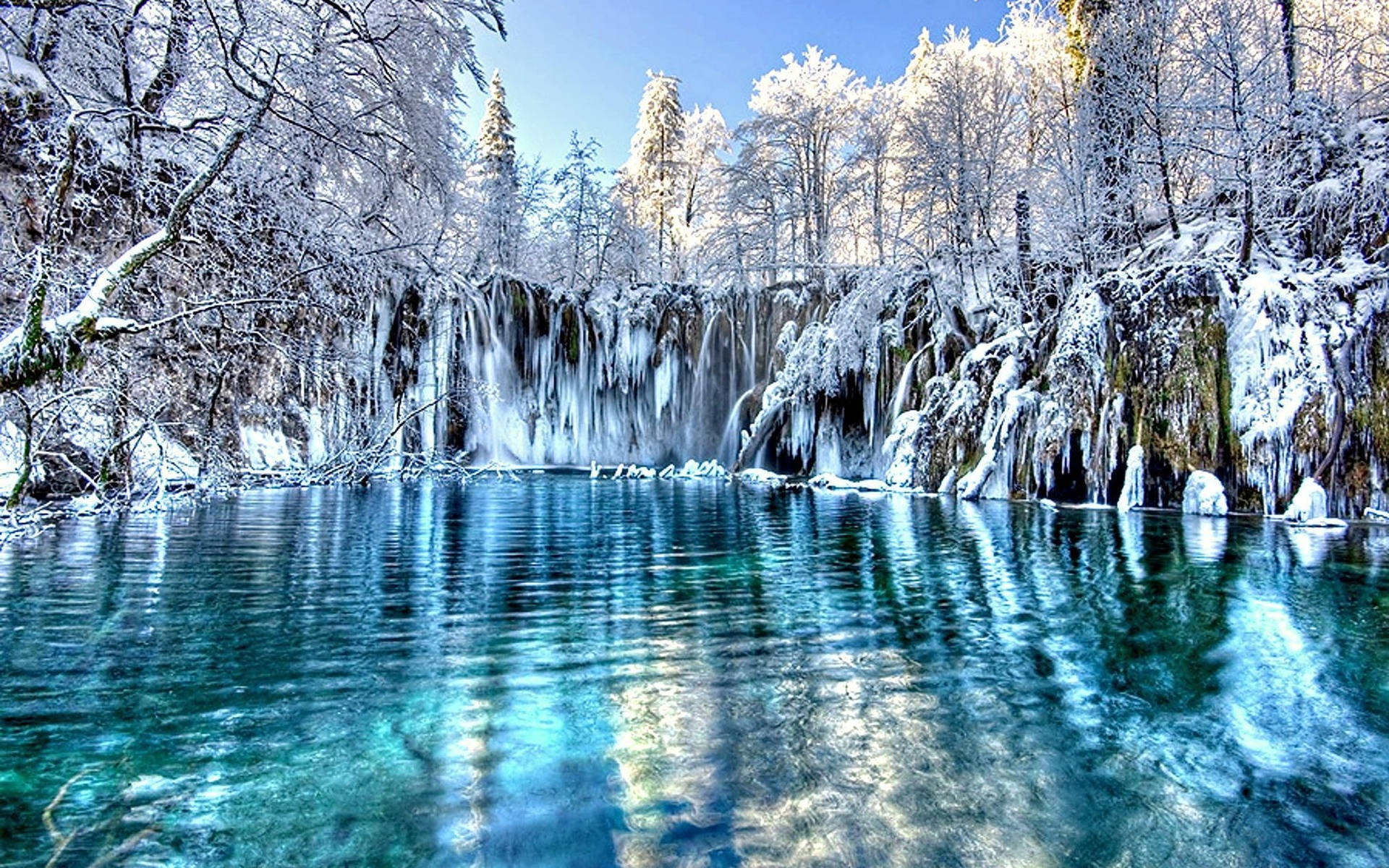雪湖森林冬景 壁紙 — 一條結冰的瀑布傾瀉而下，流入一個清澈碧綠的水潭，四周被白雪覆蓋的樹木環繞，上方是明亮的藍天。
