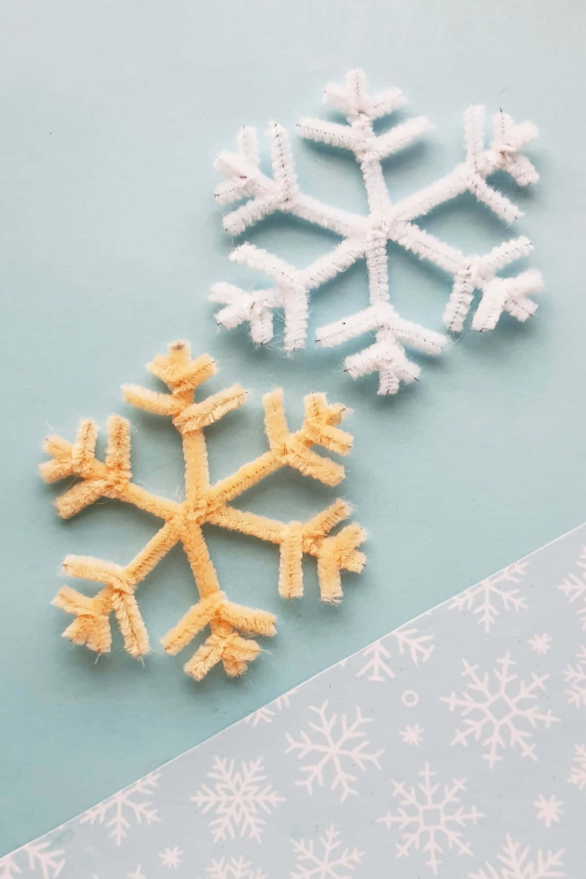 雪花圖案 壁紙 — 兩個手工製作的管道清潔器雪花——一個白色和一個淡黃色——放在柔軟的藍色表面上，旁邊是一張帶有雪花圖案的紙。