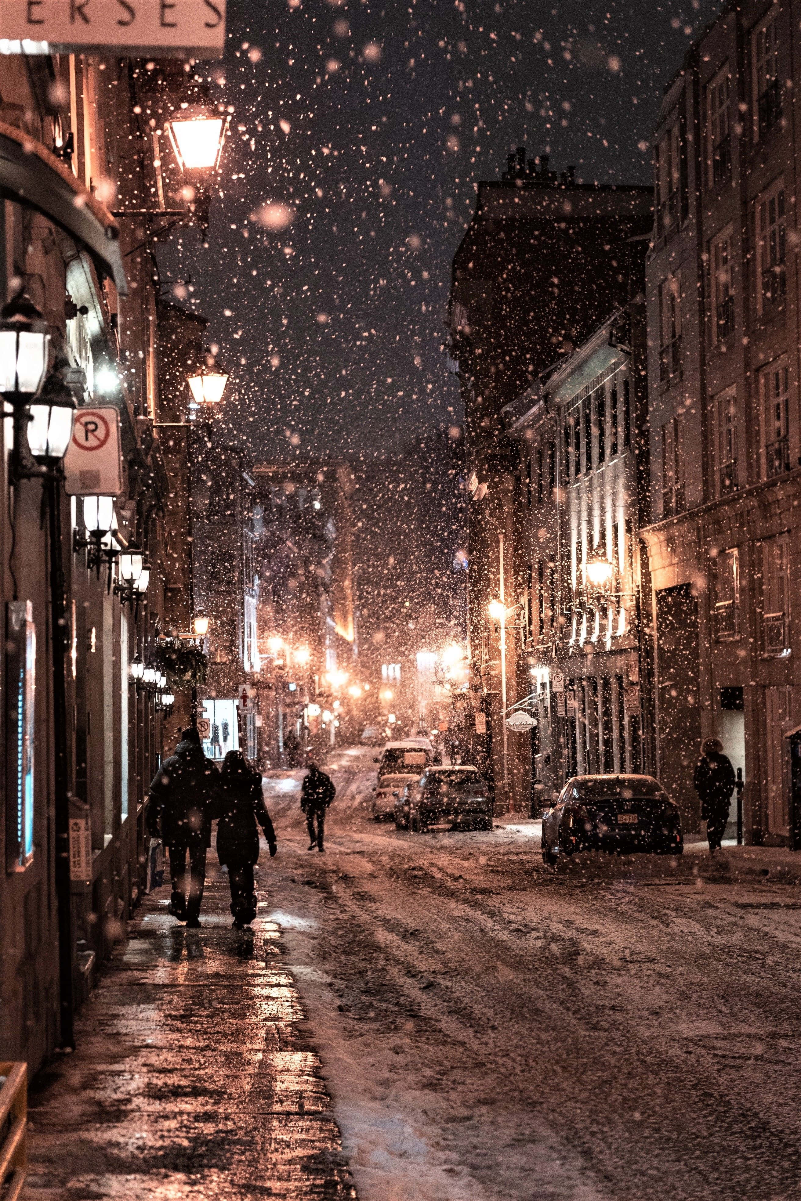 Winter Photography 桌布 壁紙 — 雪花在溫暖的路燈中飄蕩，照亮了夜晚寧靜的積雪城市小巷，有行人走過。