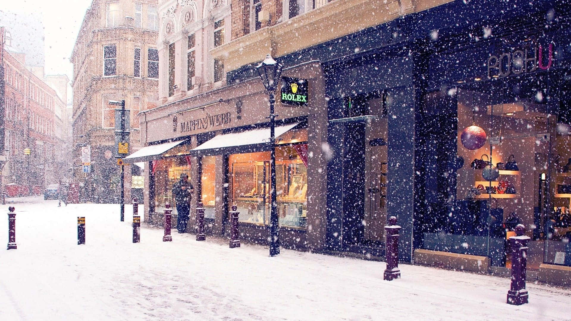 Snowy Village 桌布 壁紙 — 雪花飄過一條寧靜的城市街道，兩旁是溫暖燈光的店舖和歷史悠久的磚建築。