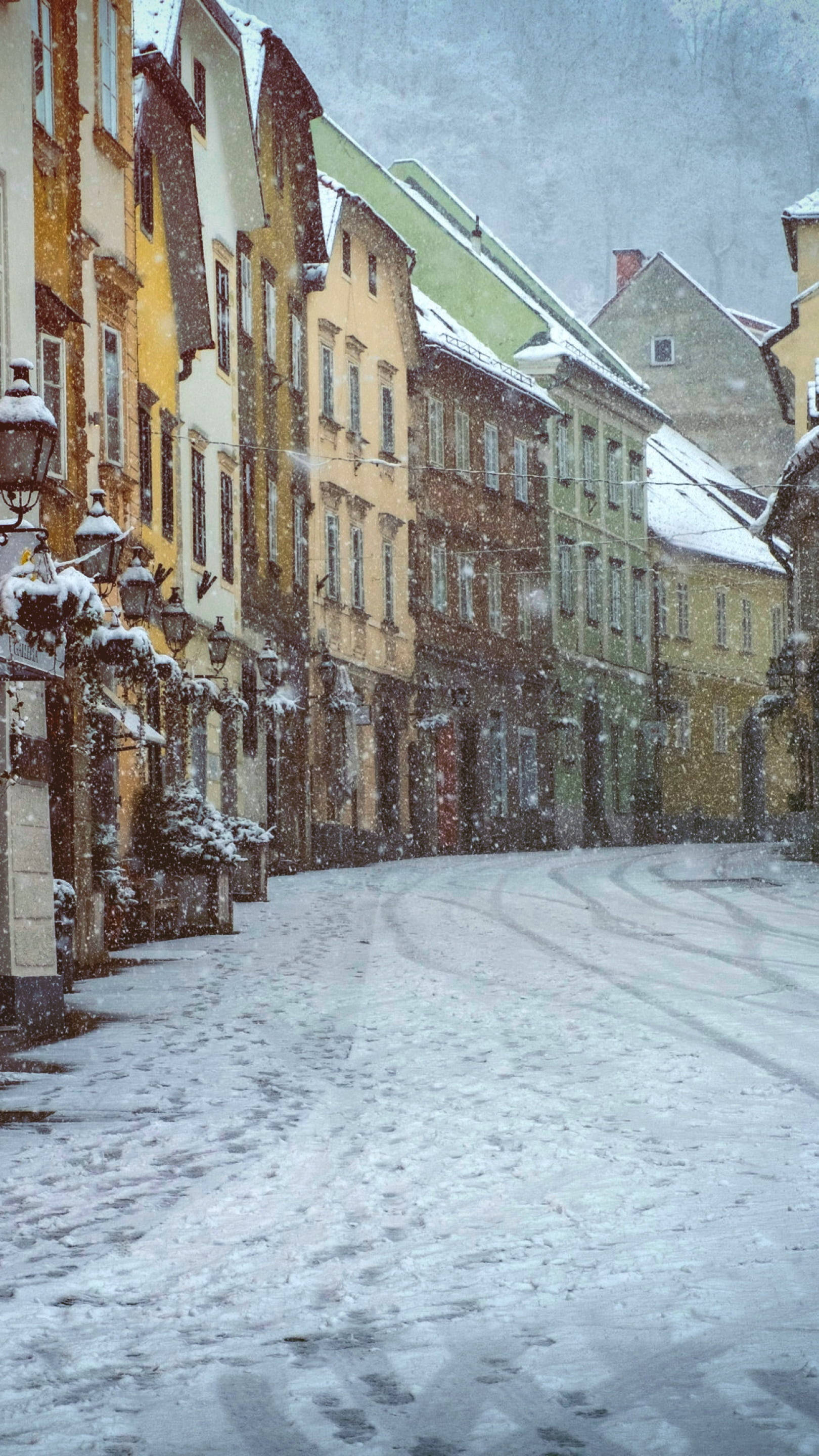 雪街彩屋溫馨冬日 壁紙 — 雪花在灰濛濛的冬日天空下，飄過一條寧靜的鵝卵石街道，兩旁是色彩繽紛的歷史建築。