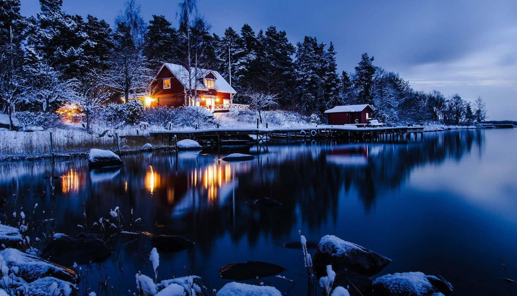 夜晚湖邊的雪中小屋，溫暖燈光倒映在平靜的水面上。