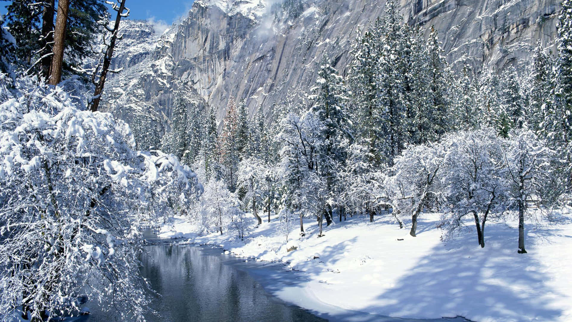 雪覆山巒與森林的冬日風景 壁紙 — 白雪覆蓋樹木和地面，旁邊是一條平靜的河流，在清澈的藍天下，靠近高聳的懸崖。