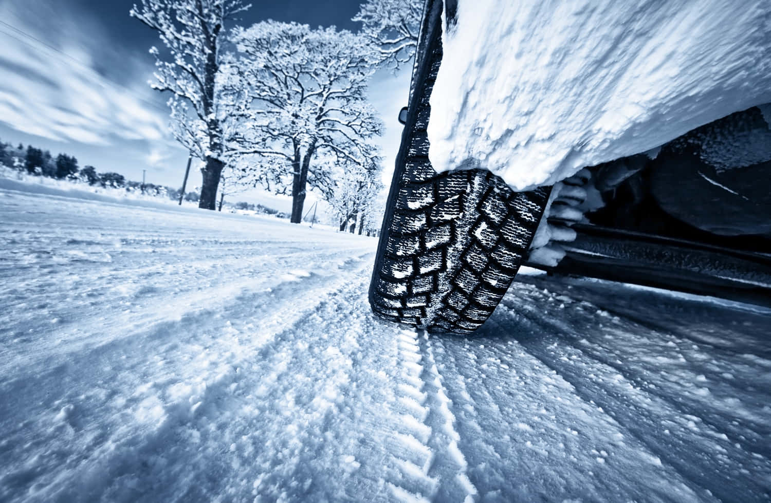 Winter Driving 桌布 壁紙 — 雪覆蓋的汽車輪胎在雪地上抓地力的特寫，背景是雪覆蓋的樹木和戲劇性的藍天。