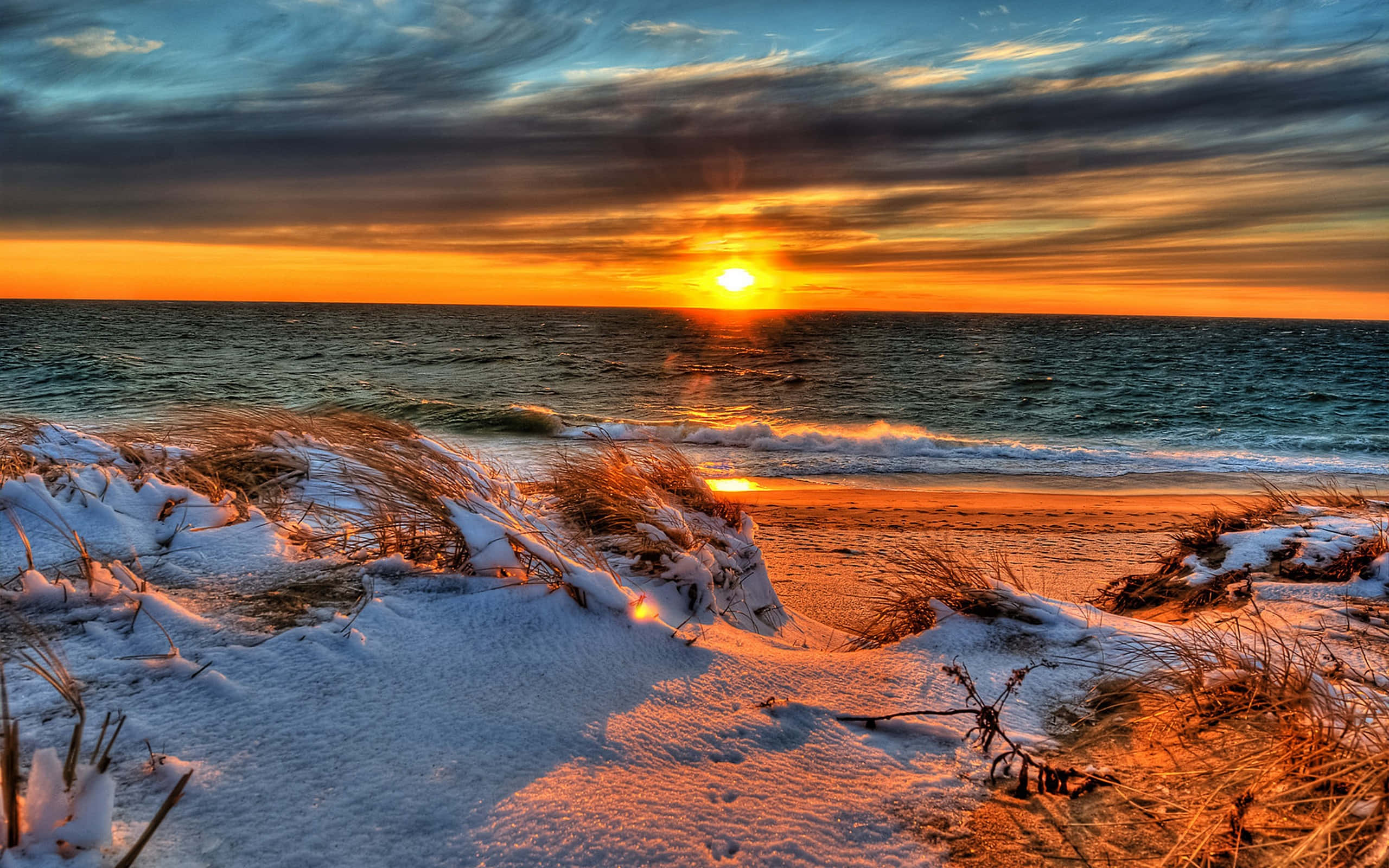Winter Beach 桌布 壁紙 — 雪覆蓋的海灘在日落時分，橙色光線反射在波浪和雪覆蓋的沙丘上，天空壯麗。