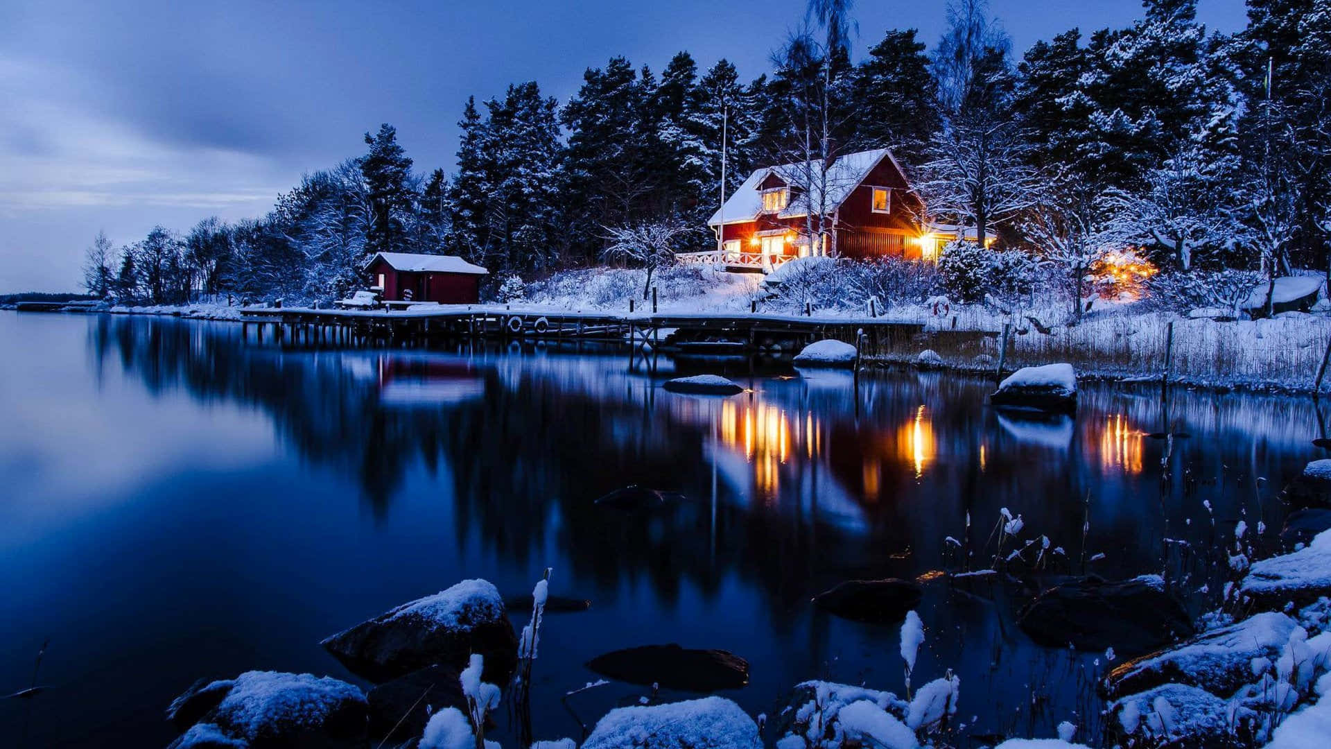 雪覆鄉野的美麗冬景 壁紙 — 一間舒適的紅色小屋，在暮色天空下，溫暖的燈光在白雪覆蓋的湖邊閃爍。