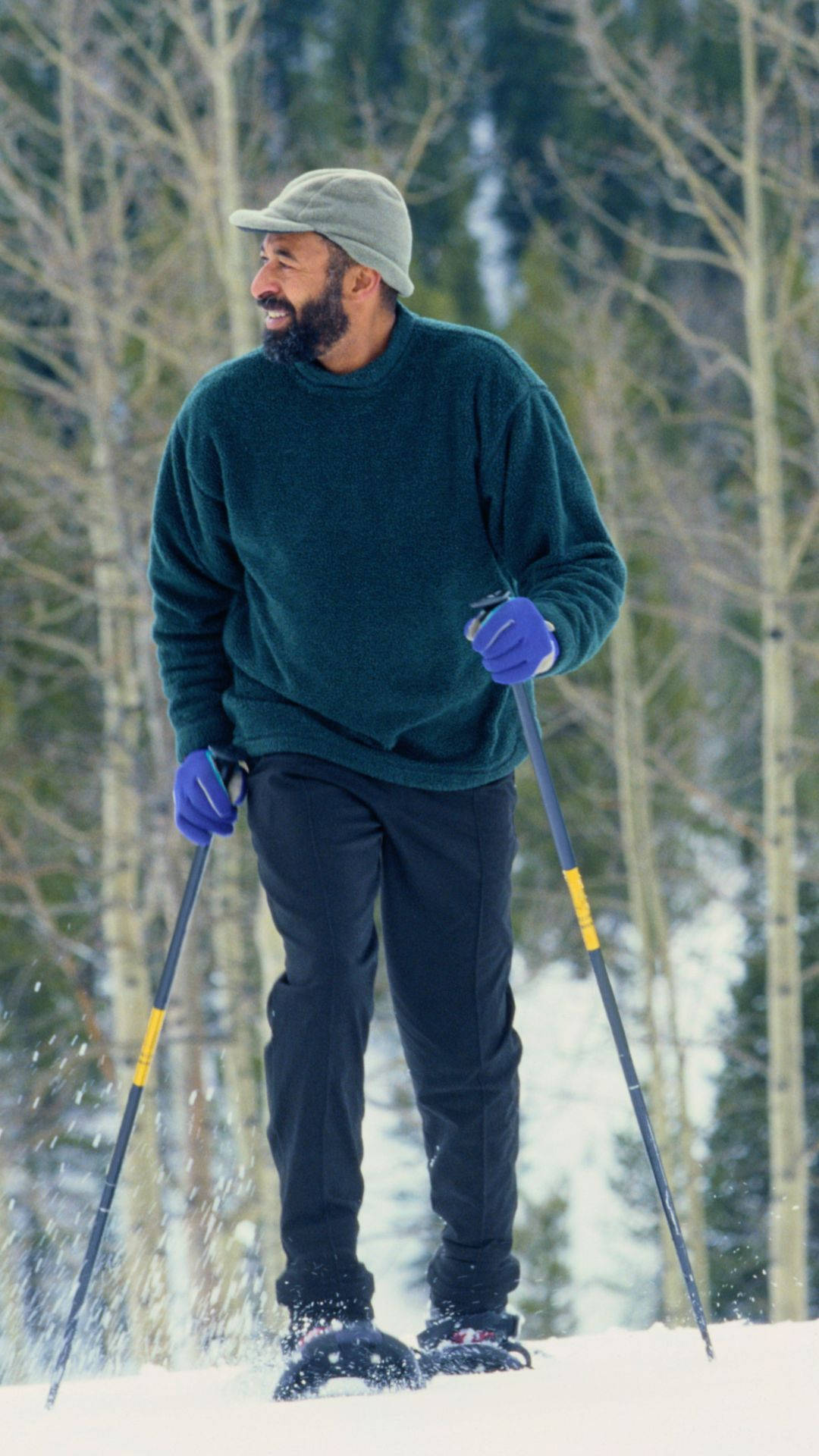 雪鞋行進偷拍照片 壁紙 — 一位留鬍鬚的男子穿著綠色毛衣和灰色帽子，在雪地森林中穿著雪鞋行走，微笑著，周圍是光禿禿的樹木。