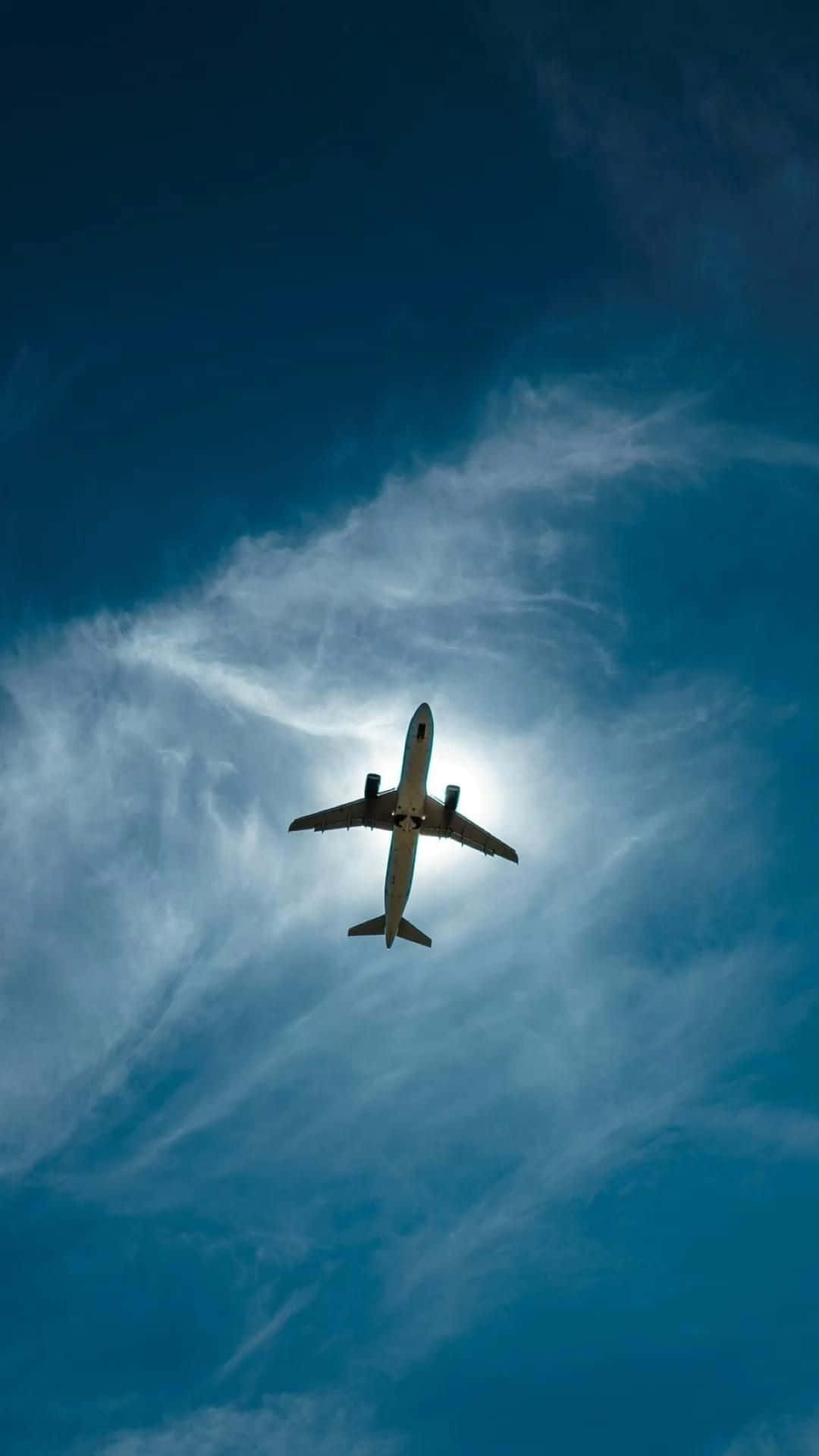 雲層天空飛機下方視角背景 壁紙 — 一架商用飛機在陽光映照下，從下方仰望，翱翔於帶有縷縷白雲的湛藍天空。