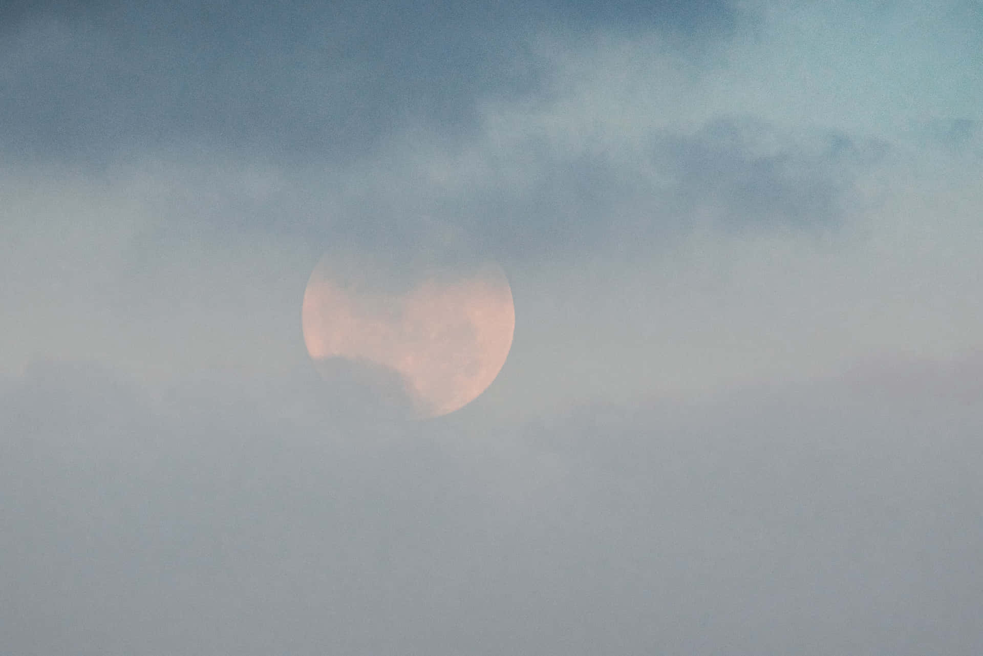 雲層後的月亮圖 壁紙 — 一輪柔和的粉紅月亮穿過縷縷灰雲，在寧靜的暮色天空中若隱若現。