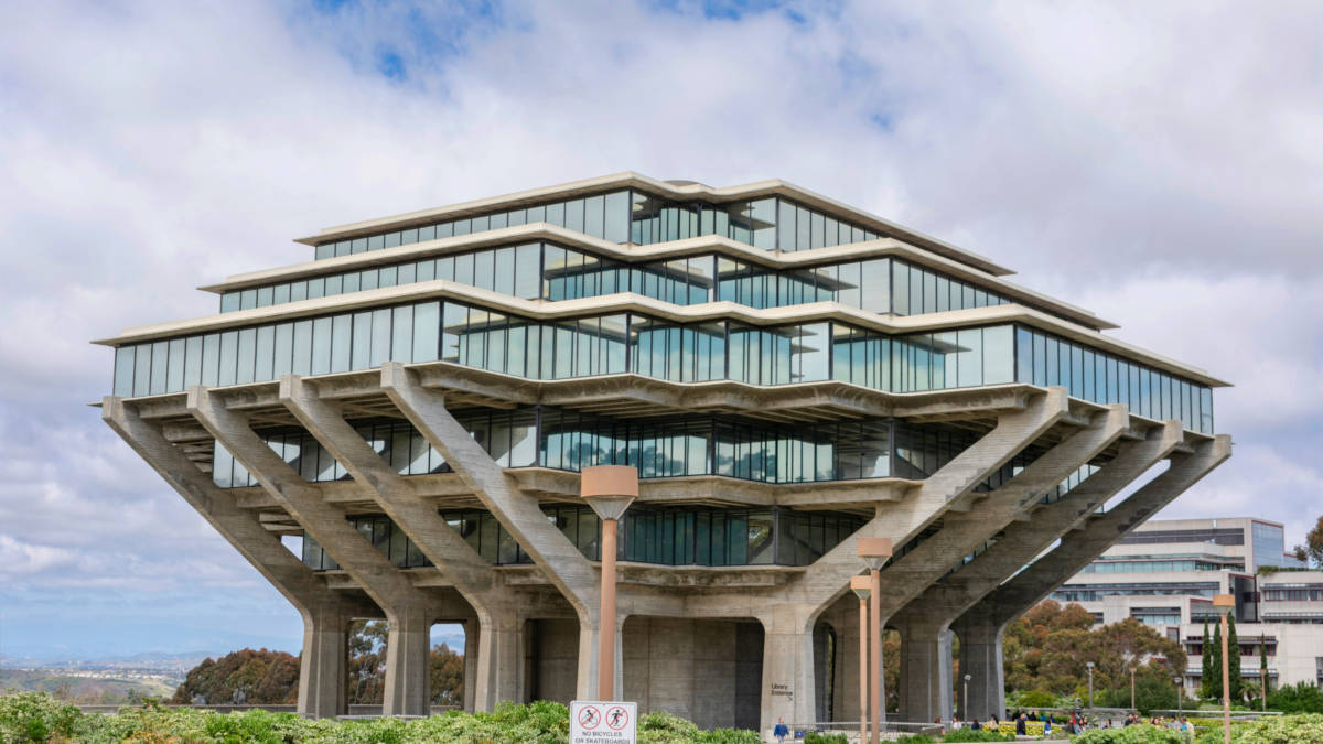 雲層籠罩下的UCSD吉賽爾圖書館 壁紙 — 一座引人注目的現代主義建築，擁有分層玻璃陽台和混凝土支撐，矗立在部分多雲的天空下。