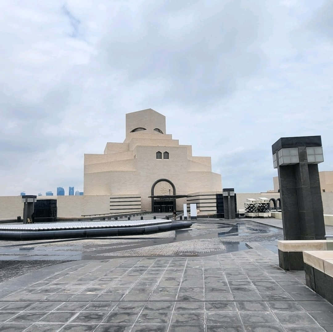 雲層籠罩下的伊斯蘭藝術博物館 壁紙 — 一座巨大的米色階梯式博物館建築，帶有拱形入口，矗立在陰天之下，潮濕的路面反射出其現代建築風格。