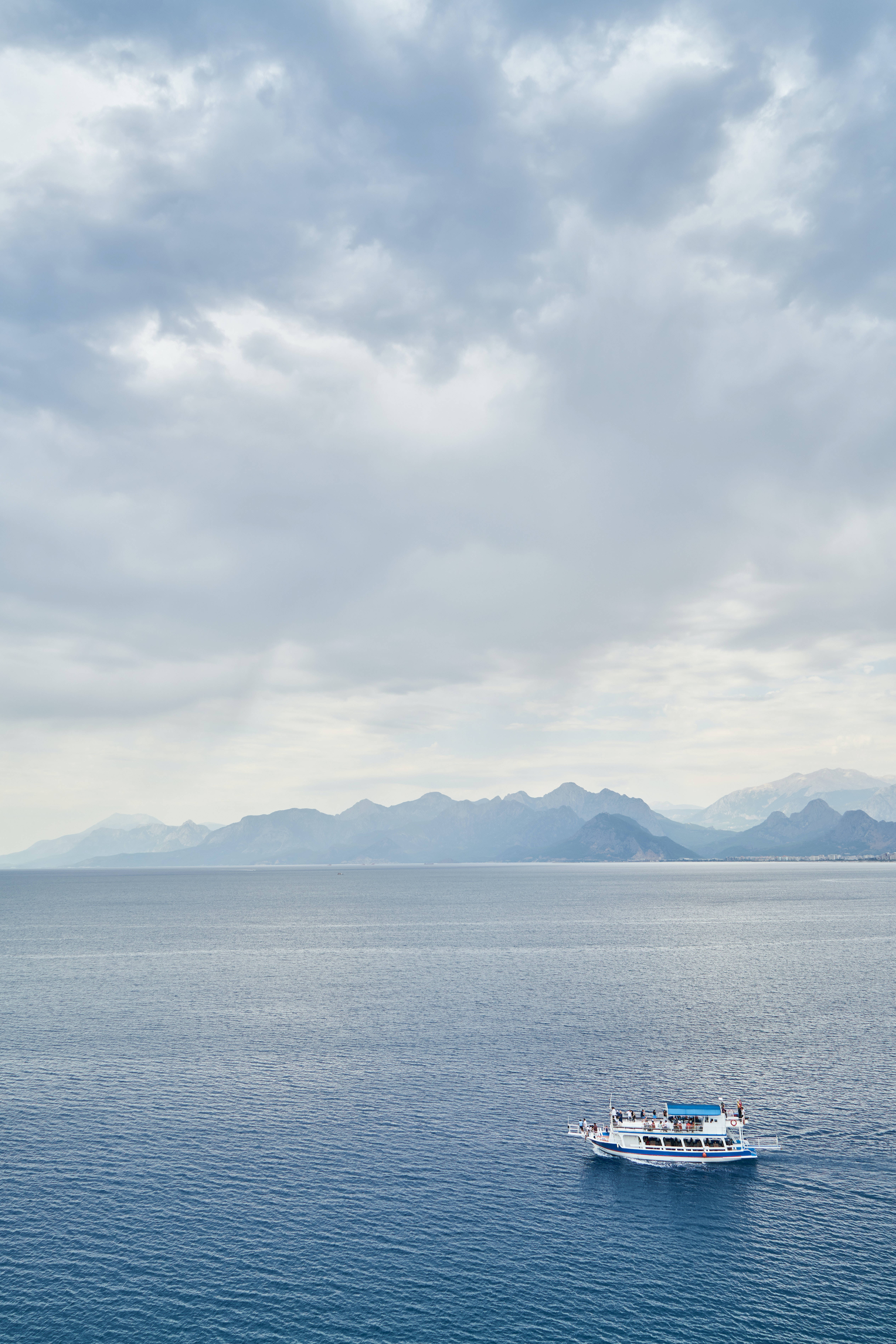 雲層覆蓋的海中船隻 壁紙 — 一艘小型藍白觀光船在深藍色水面上平靜航行，遠處山脈在陰天天空下。