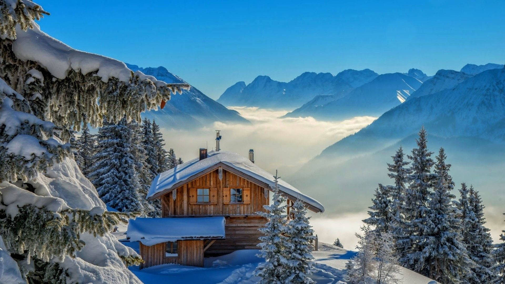雲海中的冬日小屋 壁紙 — 一間舒適的木屋坐落在白雪覆蓋的松樹林中，霧氣繚繞的山巒在雲海之上聳立，天空清澈湛藍。