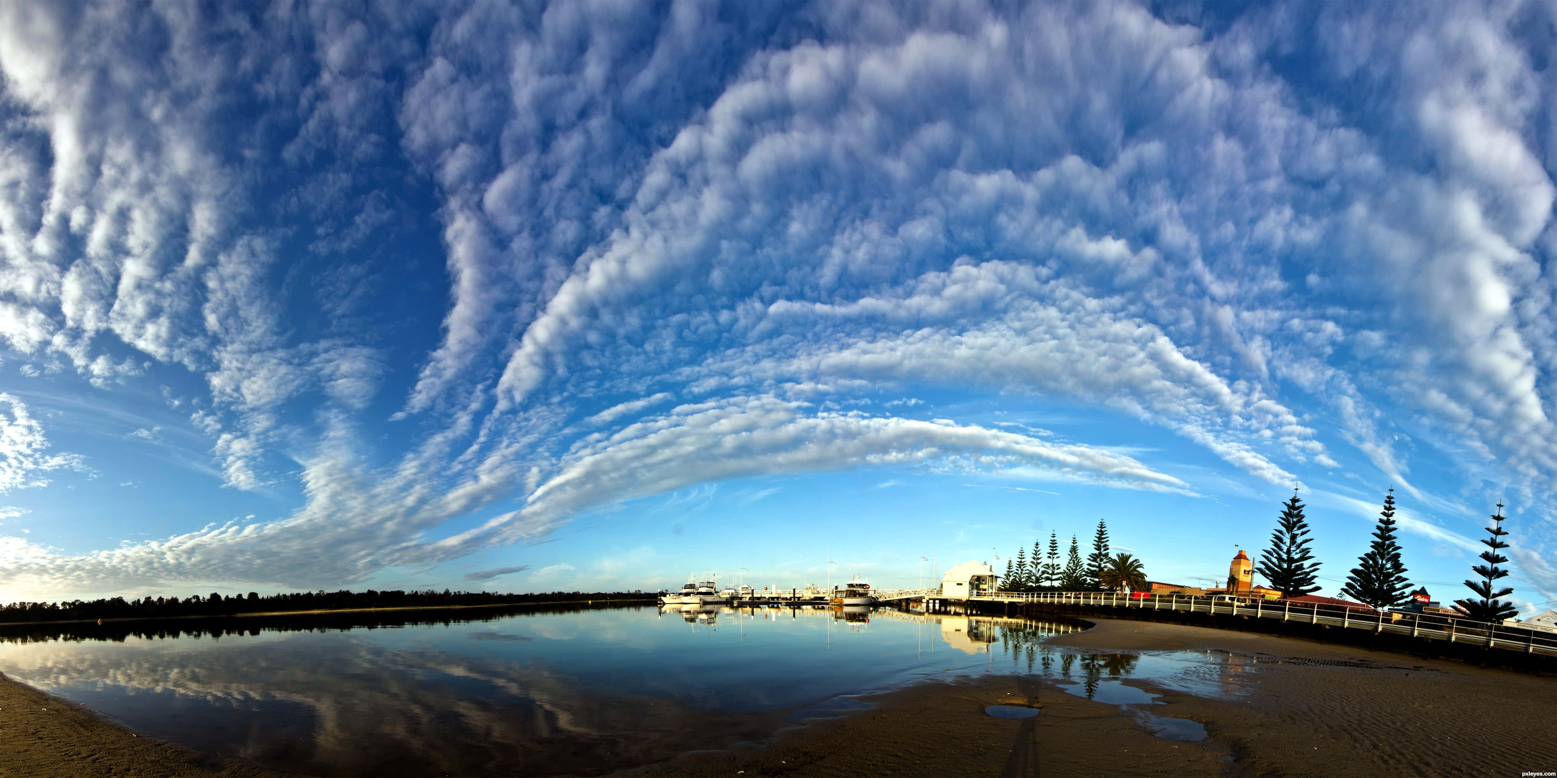 雲霧湖景 360度全景圖 壁紙 — 魚眼視角捕捉到戲劇性的白雲劃過鮮豔的藍天，下方是平靜的海濱，有船隻和松樹。