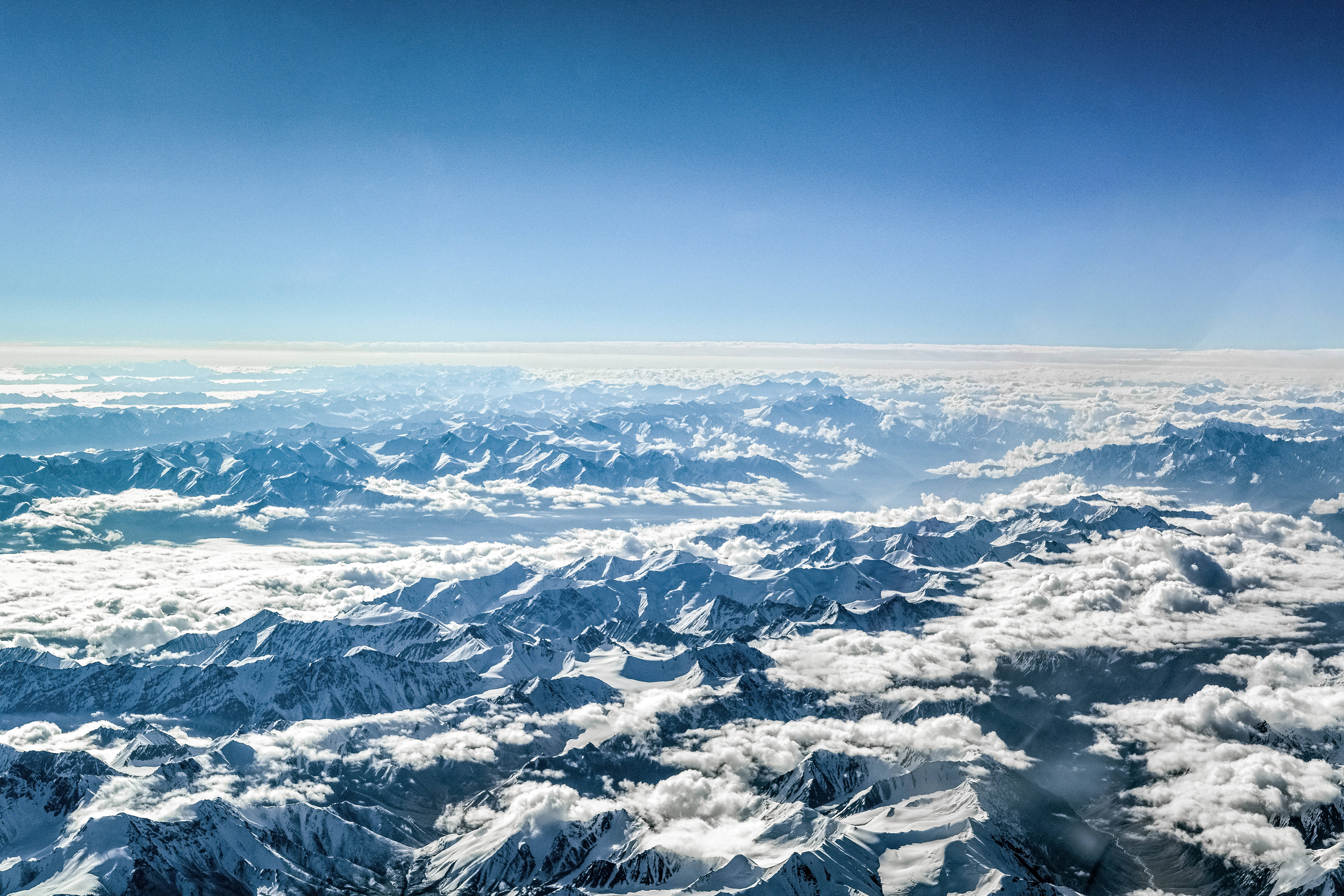 雲霧籠罩山嶺地貌 壁紙 — 從高空俯瞰，白雪覆蓋的山峰在清澈藍天下穿過蓬鬆的白雲。