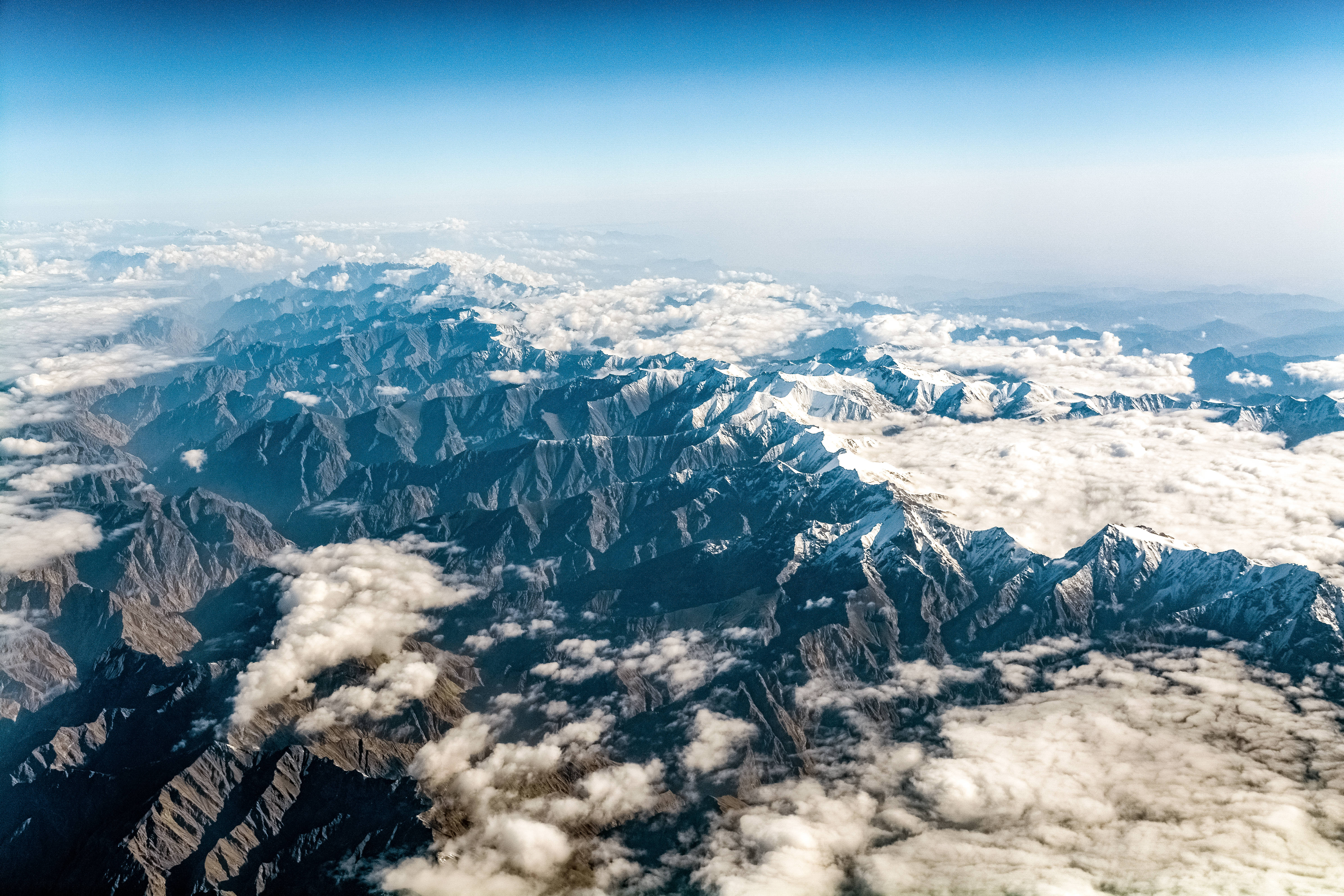 雲霧籠罩山嶺地貌 壁紙 — 從高空俯瞰，鋸齒狀的積雪山峰在清澈藍天下穿過蓬鬆的白雲。
