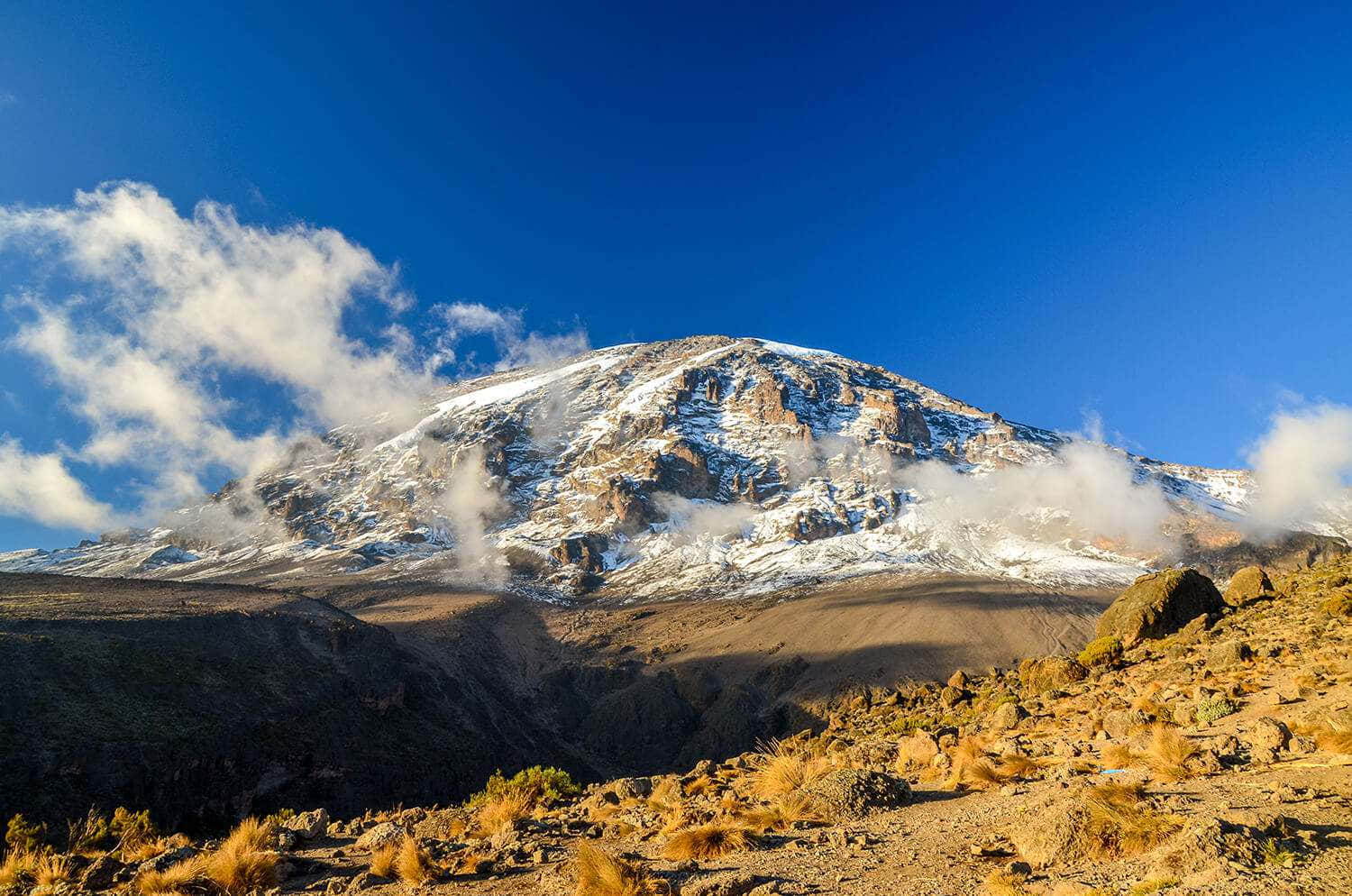 在清澈的藍天下，雲朵環繞著岩石山坡的雪山峰頂。