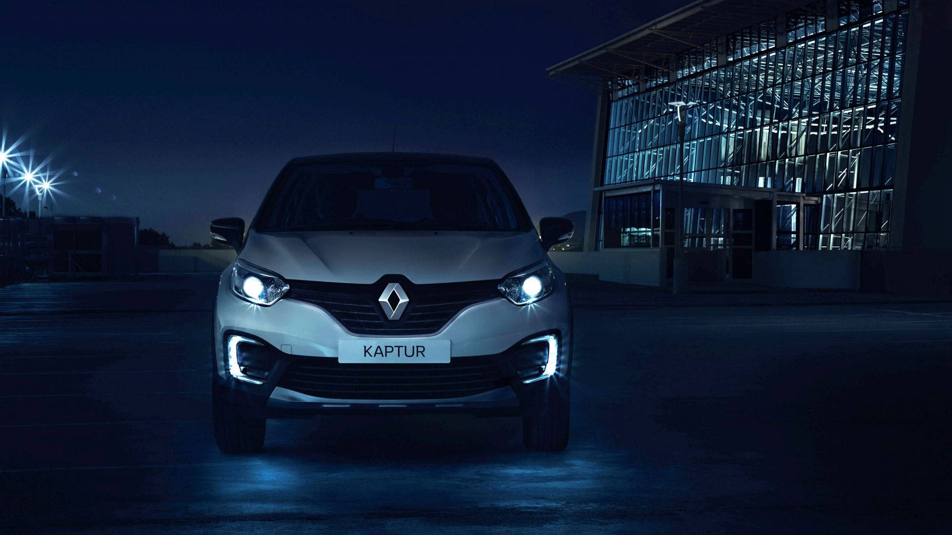 銀色雷諾Captur SUV在夜晚被車頭燈照亮，停在一棟現代玻璃建築前。