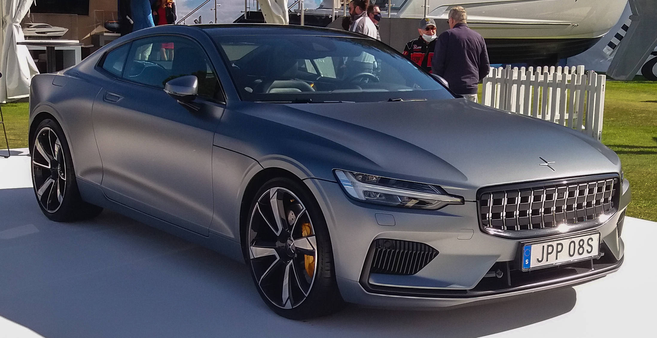 雷霆色 Polestar 1 壁紙 — 一輛時尚的啞光銀色跑車，配備黑色輪圈和黃色制動卡鉗，在戶外活動的白色平台上展示。