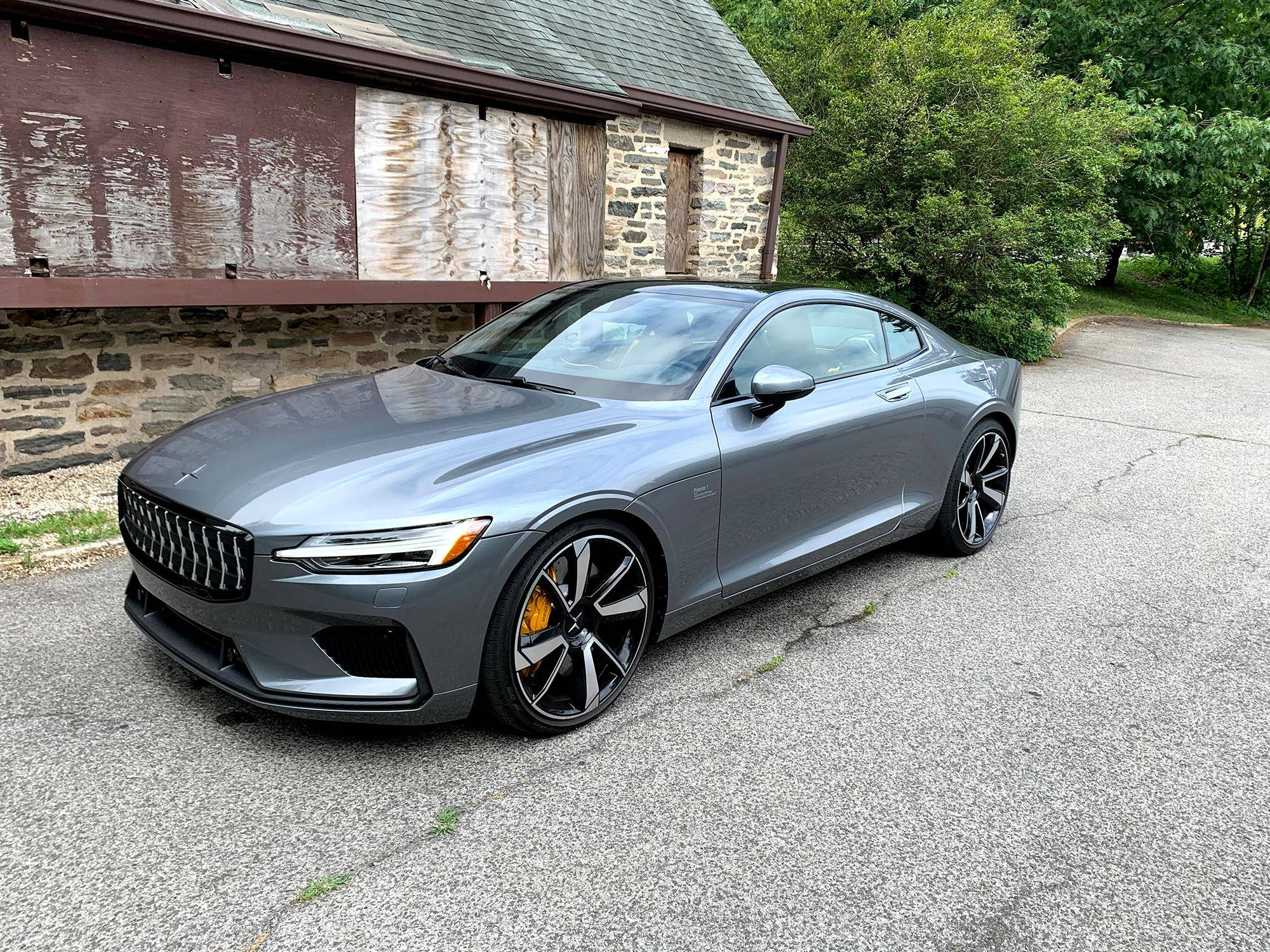 雷霆色 Polestar 電動車 壁紙 — 一輛時尚的灰色Polestar 2電動轎跑車停在裂開的瀝青車道上，旁邊是一座質樸的石木建築，背景是茂密的綠樹。