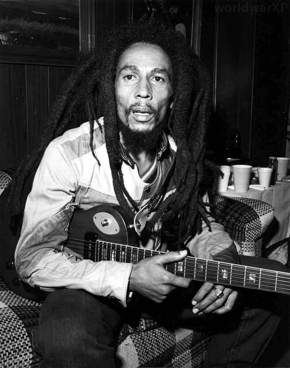 雷鬼之王鮑勃·馬利 壁紙 — Bob Marley 坐喺昏暗嘅房入面揸住結他，背景有圖案傢俬同杯。