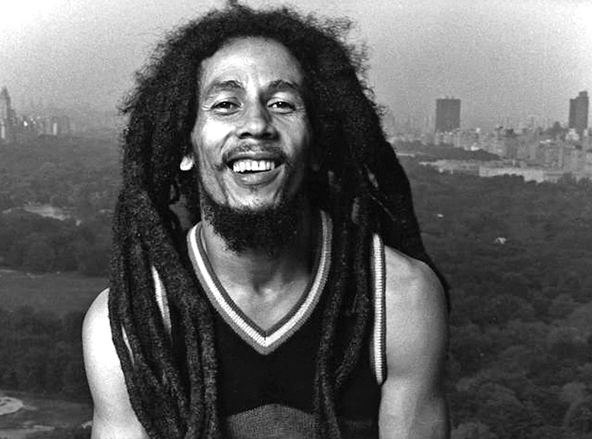 雷鬼音樂的傳奇與先驅——鮑勃·馬利 壁紙 — Bob Marley 溫暖地微笑，長髮綹框住他的臉龐，背景是朦朧的紐約天際線和下方的綠色公園。