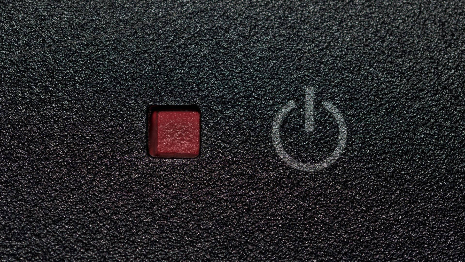 電力中斷時的黑色UPS開關 壁紙 — 紅色電源按鈕在帶有紋理的黑色表面上發光，帶有白色電源符號的特寫。
