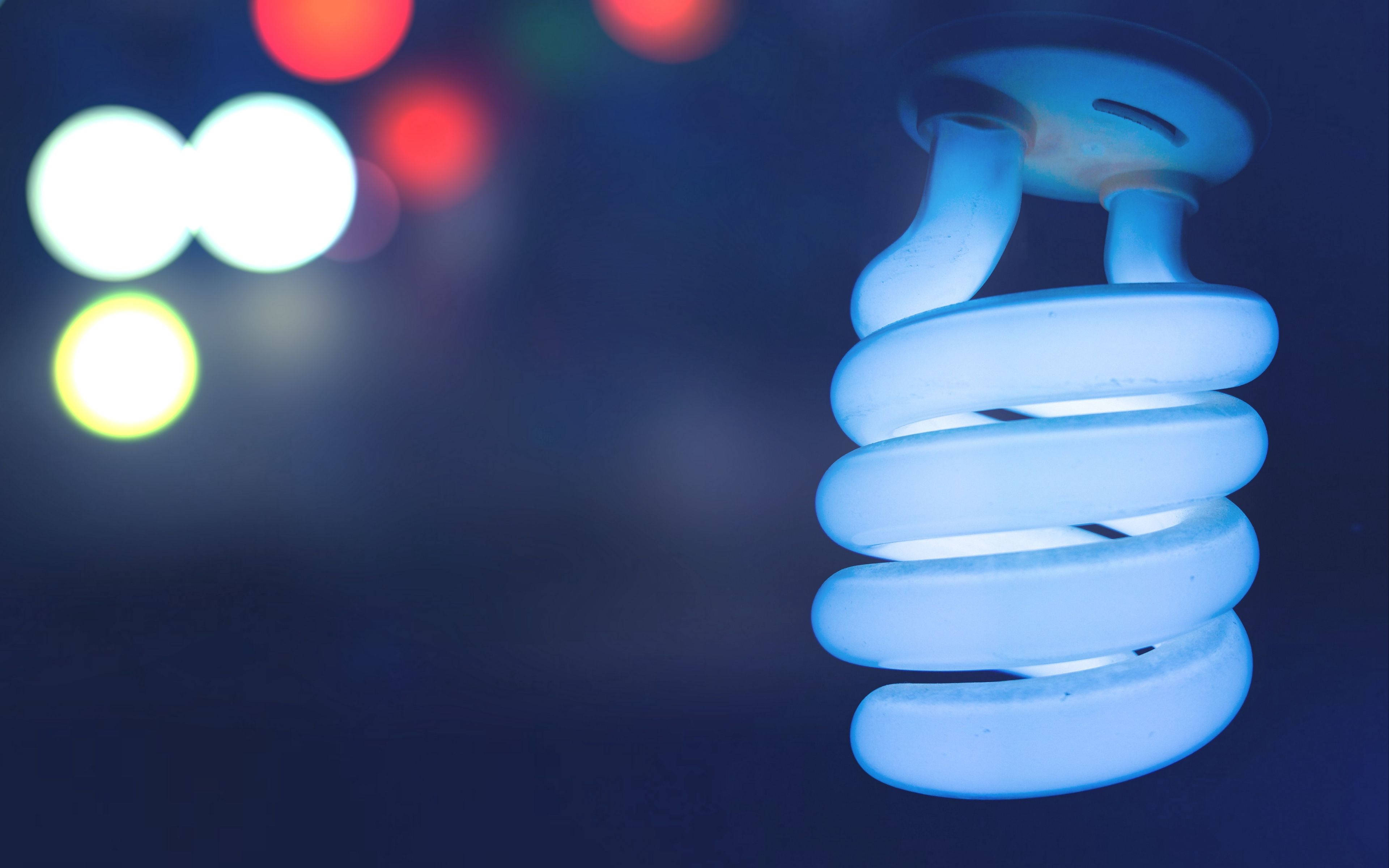 電力燈泡散景 壁紙 — 一個發光的藍色緊湊型熒光燈泡懸掛在黑暗背景上，遠處有彩色模糊燈光。