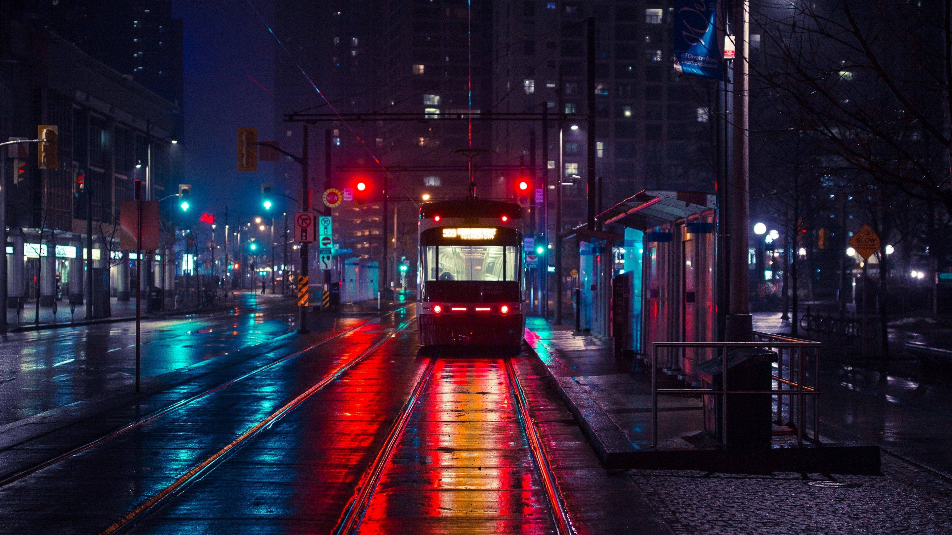 電力驅動電車 壁紙 — 一輛紅色電車在雨濕的城市夜晚中滑行，其燈光在高聳建築物下的濕滑路軌和路面上生動地反射。